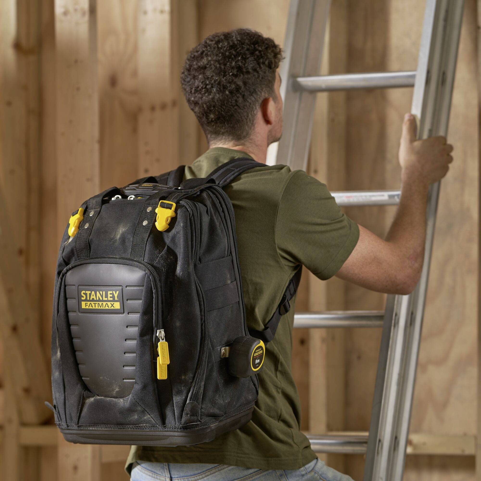 Ein Mann steht auf einer Leiter und trägt einen schwarzen Stanley Fatmax-Rucksack. Er ist in einer Werkstattumgebung.
