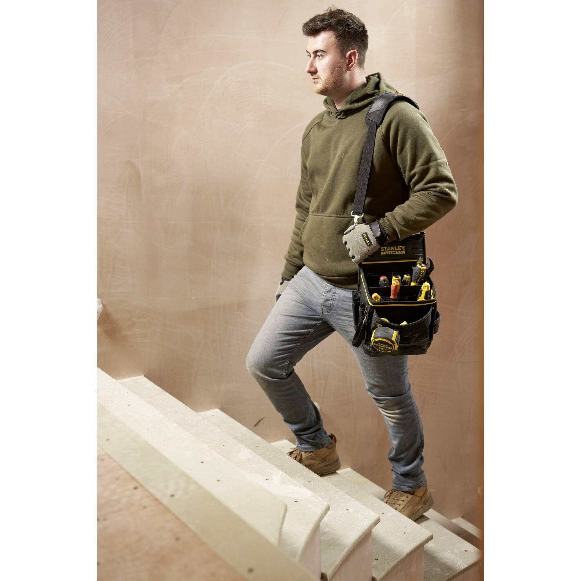 Ein Handwerker in Freizeitkleidung steigt eine Treppe hinauf. Er trägt eine Werkzeugtasche über der Schulter und Handschuhe.