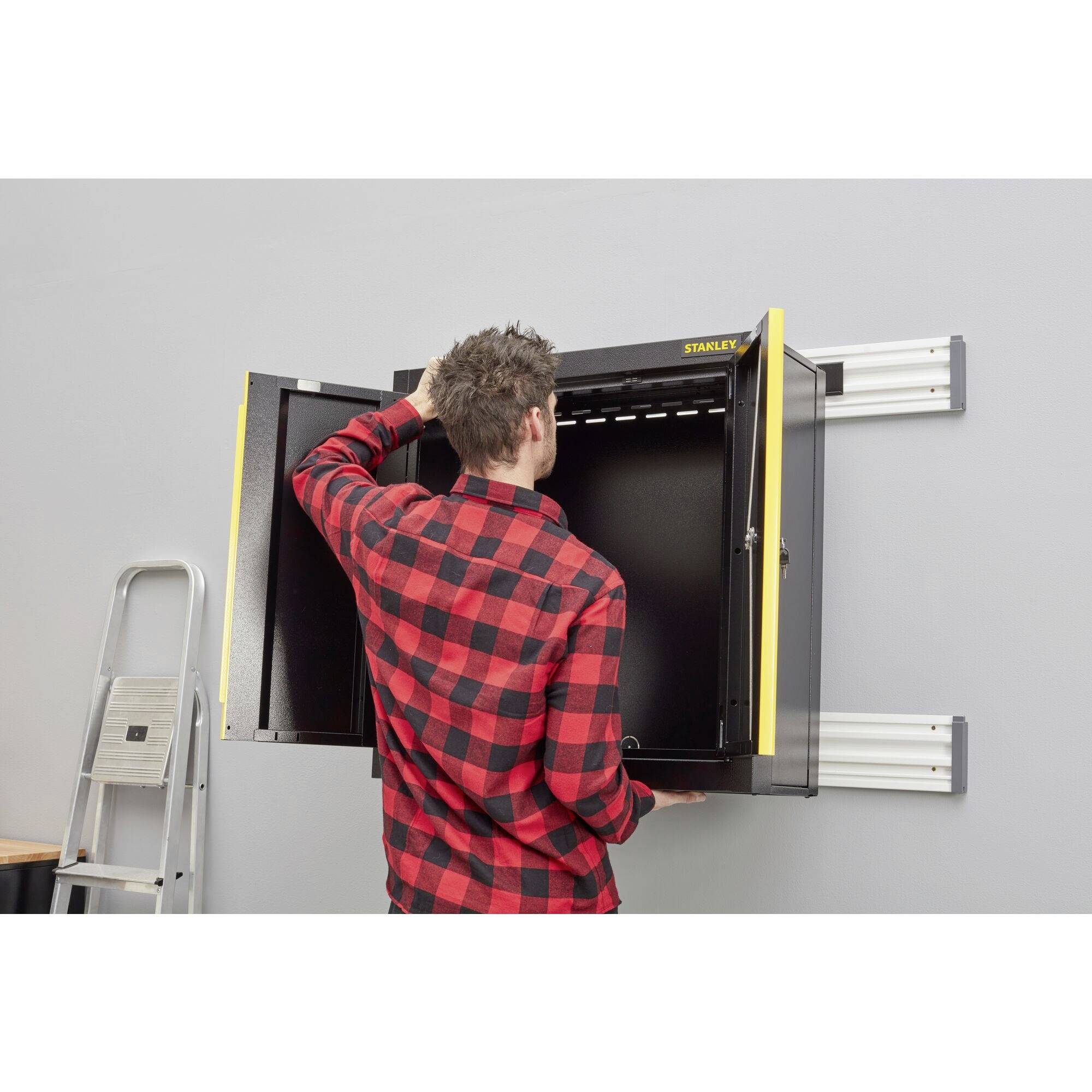 Ein Mann öffnet einen schwarzen Wandschrank an einer grauen Wand. Eine Leiter steht seitlich, der Raum wirkt organisiert und funktional.