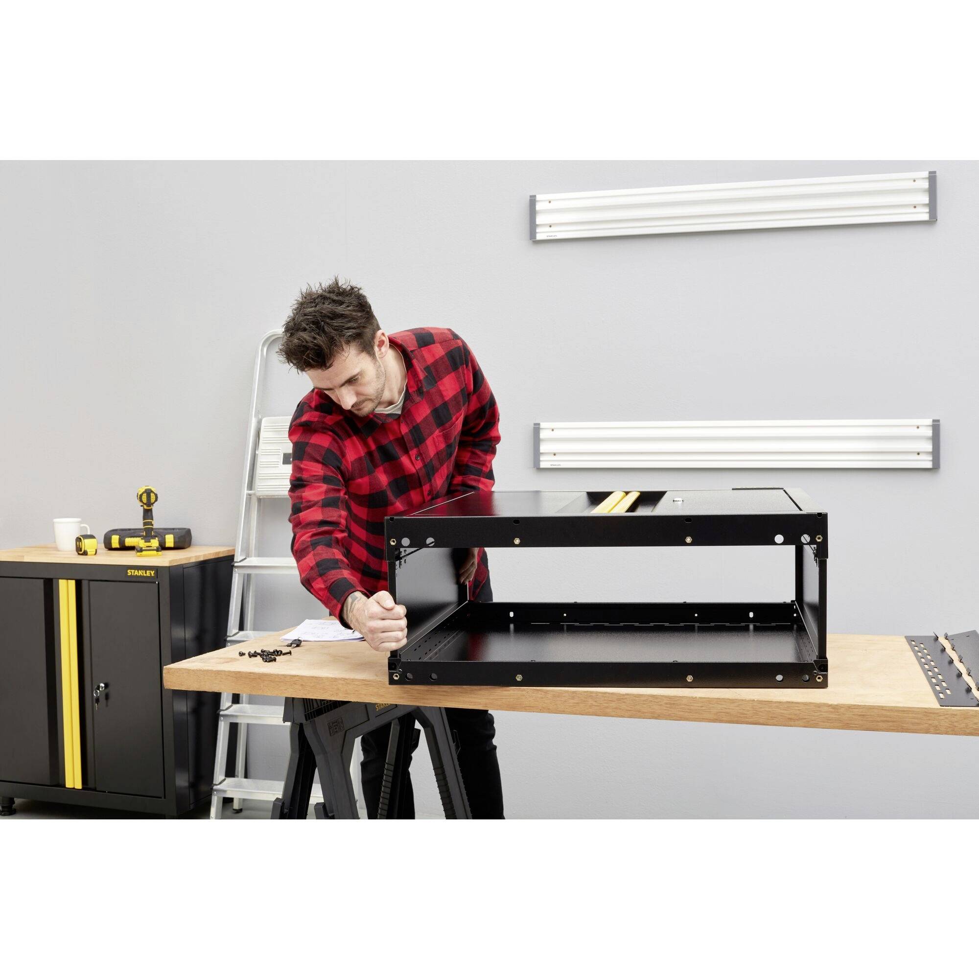 Ein Mann in kariertem Hemd montiert ein schwarzes Regal in einer Werkstatt. Werkzeugschrank und Leiter im Hintergrund.