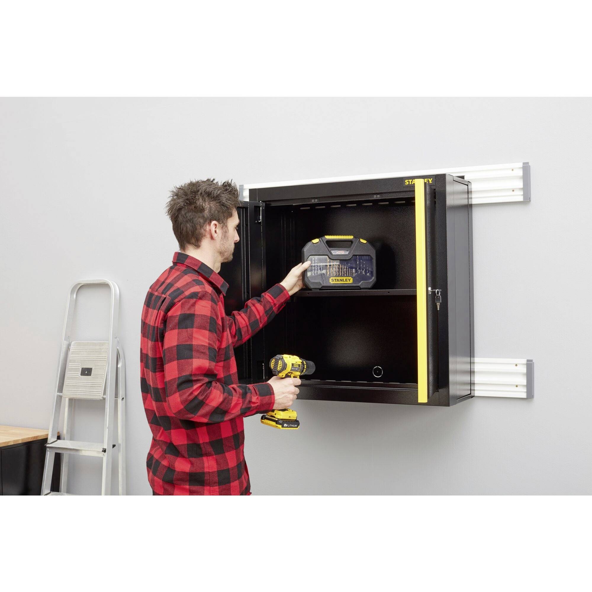 Ein Mann in einem karierte Hemd steht vor einem geöffneten schwarzen Wandschrank, hält eine Akku-Bohrmaschine und eine Werkzeugbox. Links steht eine Leiter.