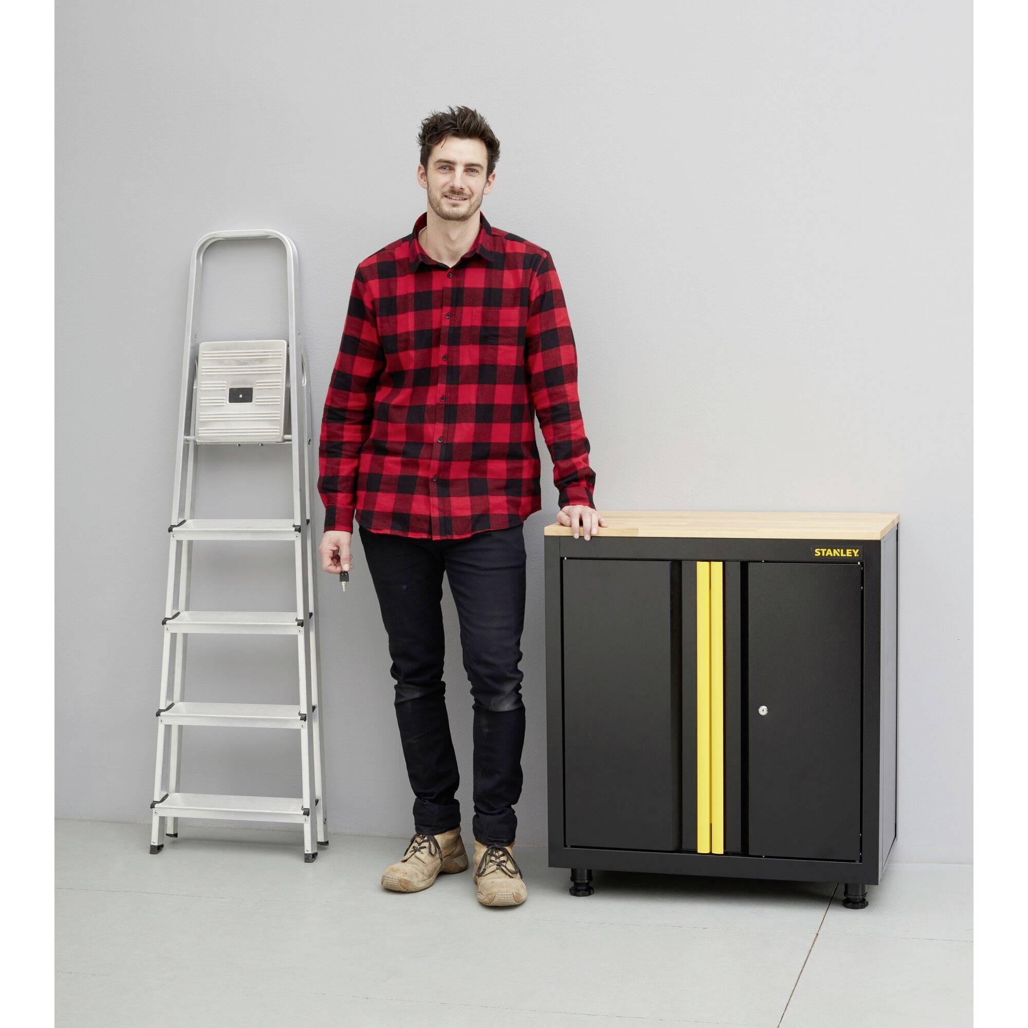 Ein Mann steht neben einem schwarzen Schrank mit Holztop und gelben Streifen; links steht eine zusammengeklappte Leiter an der Wand.