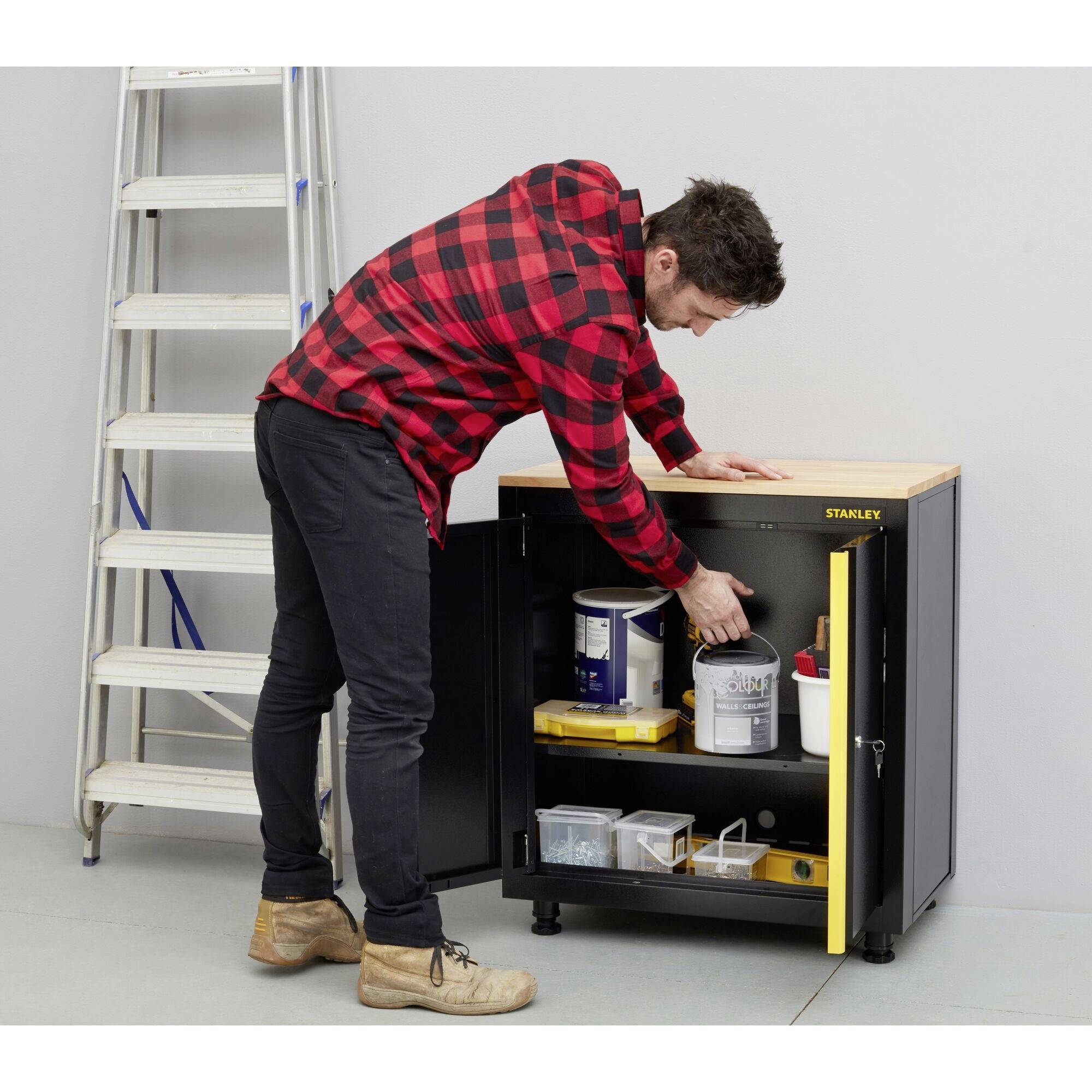 Ein Mann in kariertem Hemd entnimmt Farbmaterialien aus einem Werkzeugschrank. Im Hintergrund steht eine Leiter.