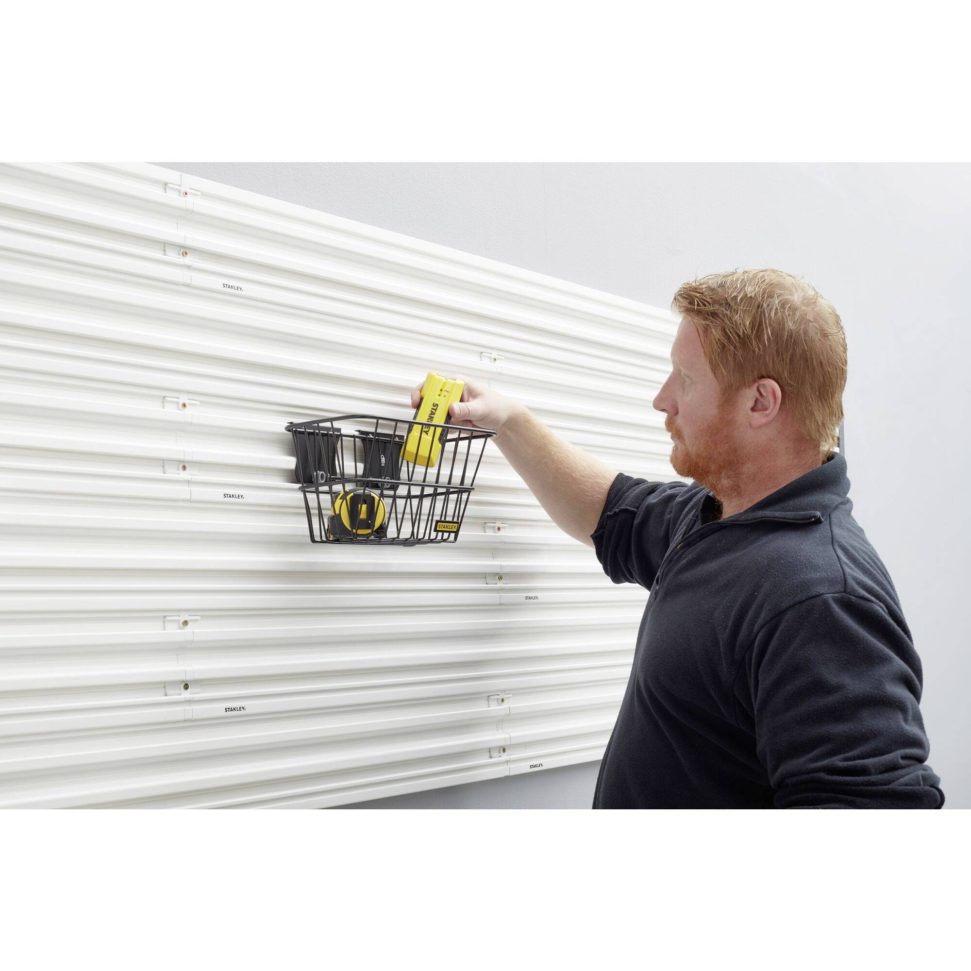 Ein Mann hängt einen schwarzen Metallkorb an eine weiße, gerillte Wand. Der Korb enthält Werkzeuge wie ein Maßband.