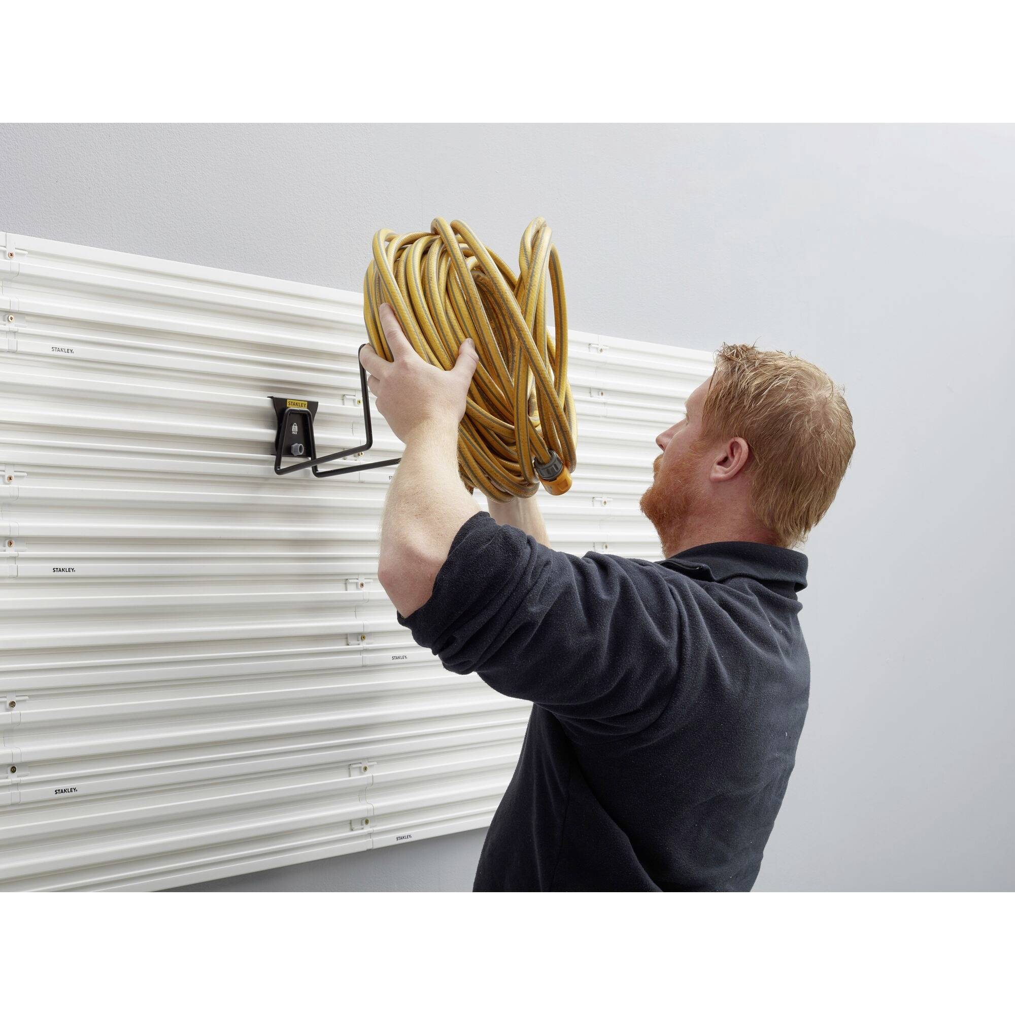 Ein Mann befestigt einen gelben Schlauch an einem Wandhaken in einer Werkstatt. Die Wand ist mit horizontalen Schienen verkleidet.