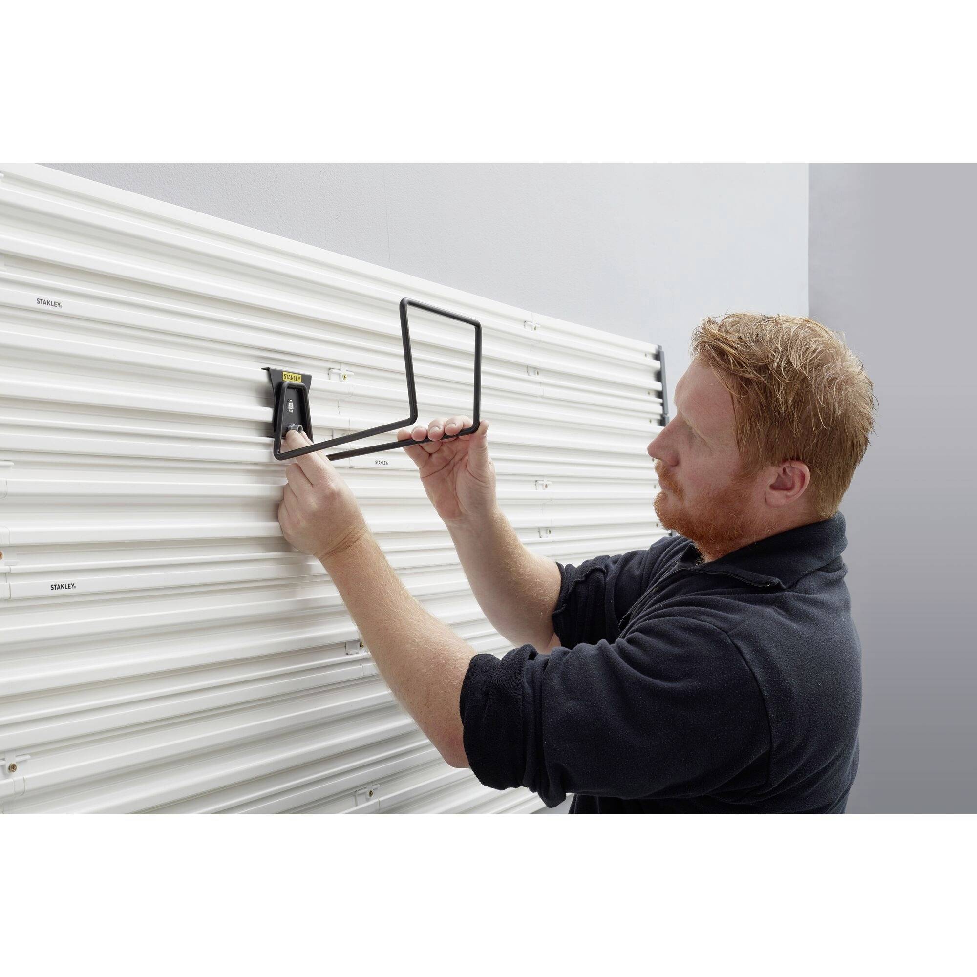 Eine Person befestigt eine schwarze Halterung an einer weißen, geriffelten Wand. Sie trägt dunkle Kleidung und arbeitet konzentriert.