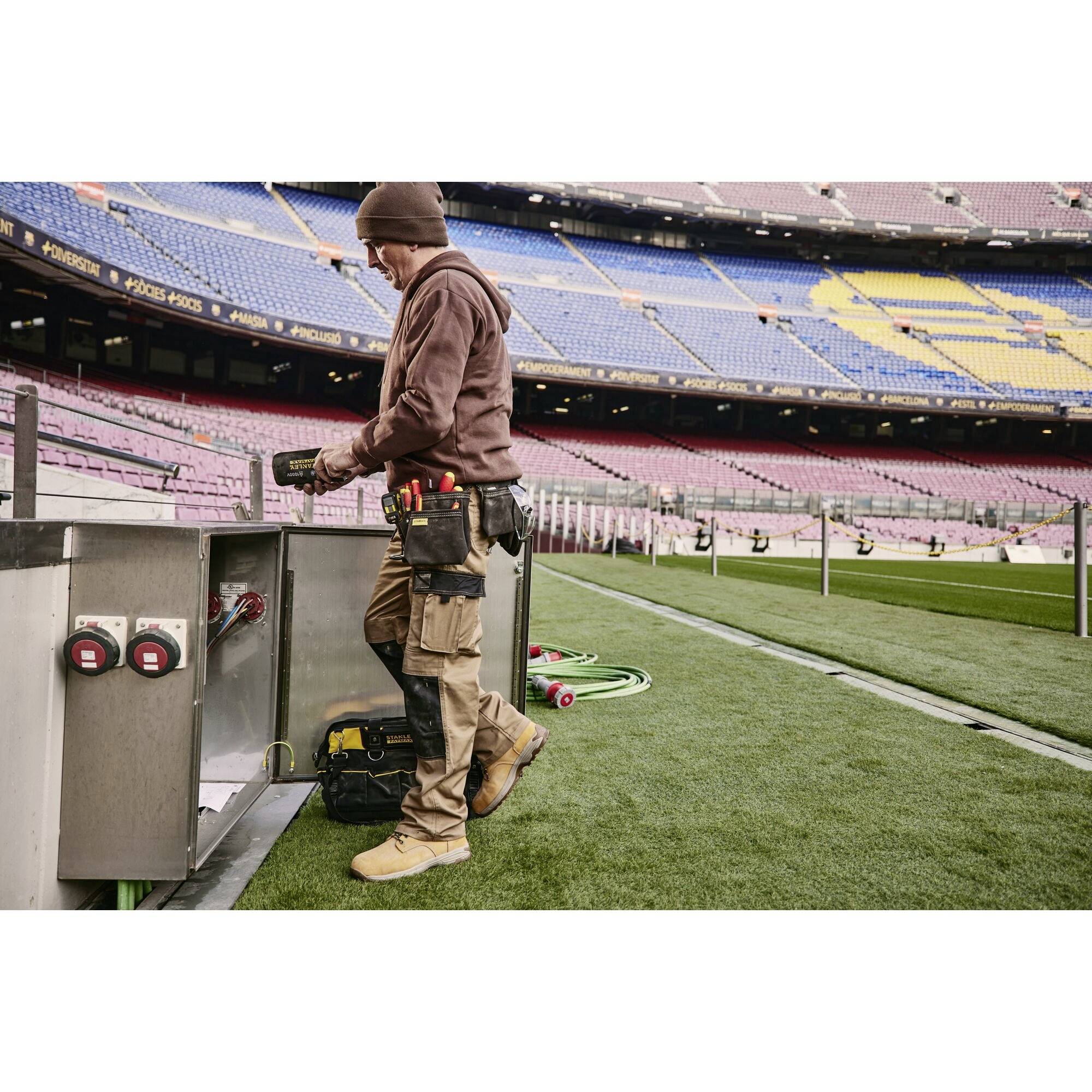 Eine Person im Arbeitsanzug steht an der Seitenlinie eines großen leeren Stadions und betrachtet ein Panel mit Wasseranschlüssen.