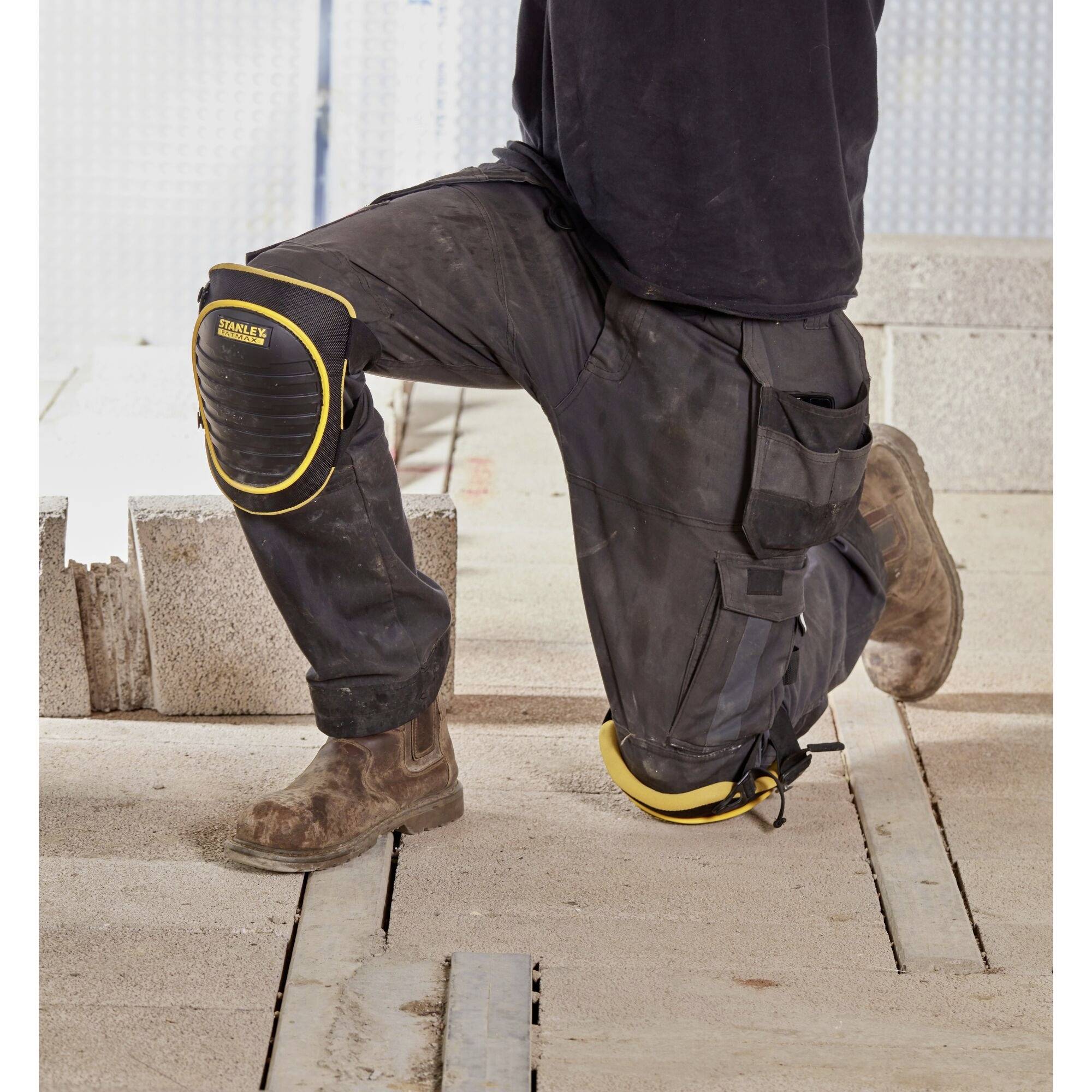 Eine Person kniet auf Baustellenmaterial, trägt knieschonende Arbeitskleidung und Sicherheitsstiefel. Betonsteine sind im Hintergrund.