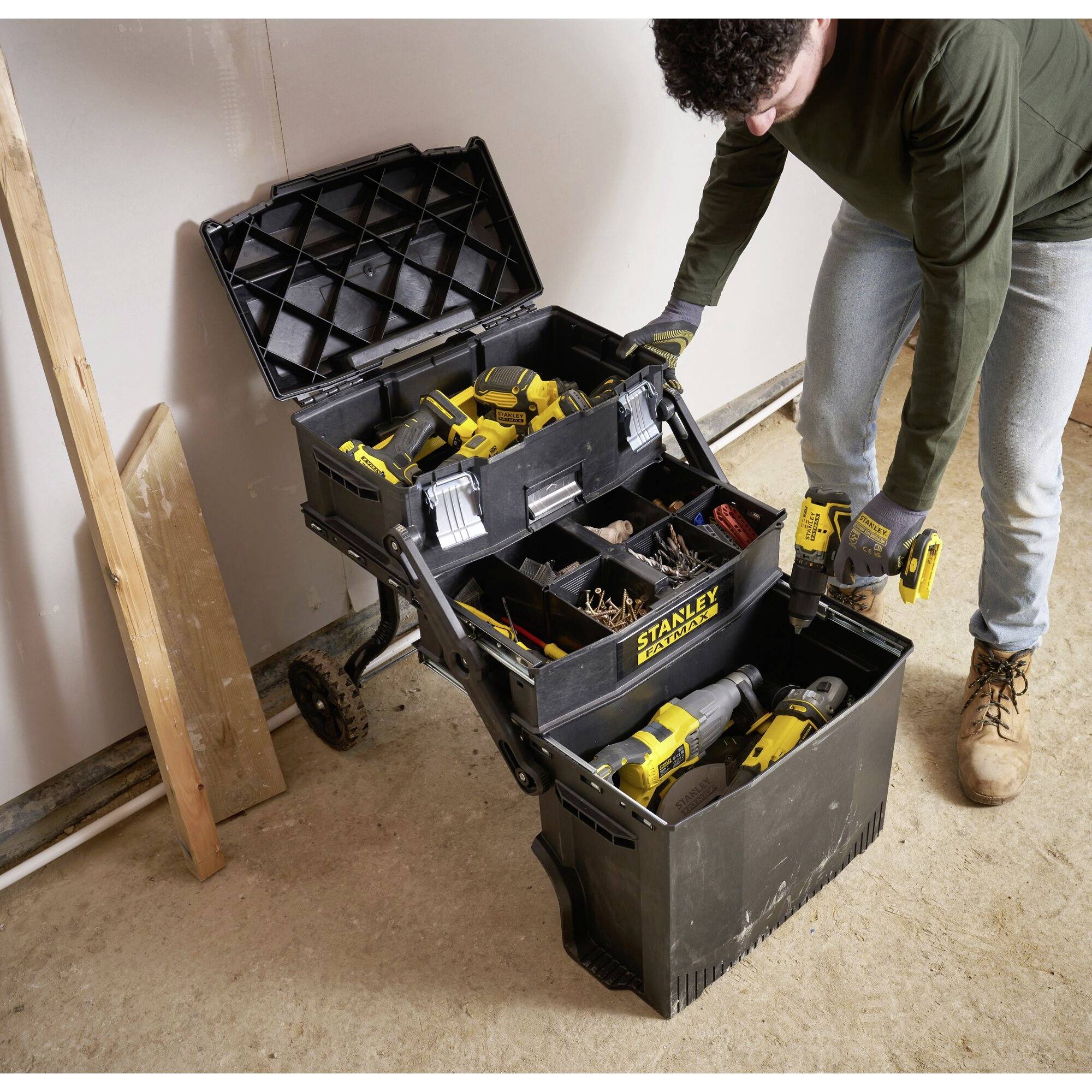 Eine Person ordnet Werkzeuge in einem schwarzen, offenen Werkzeugkasten mit mehreren Fächern auf Rädern in einer Werkstatt.