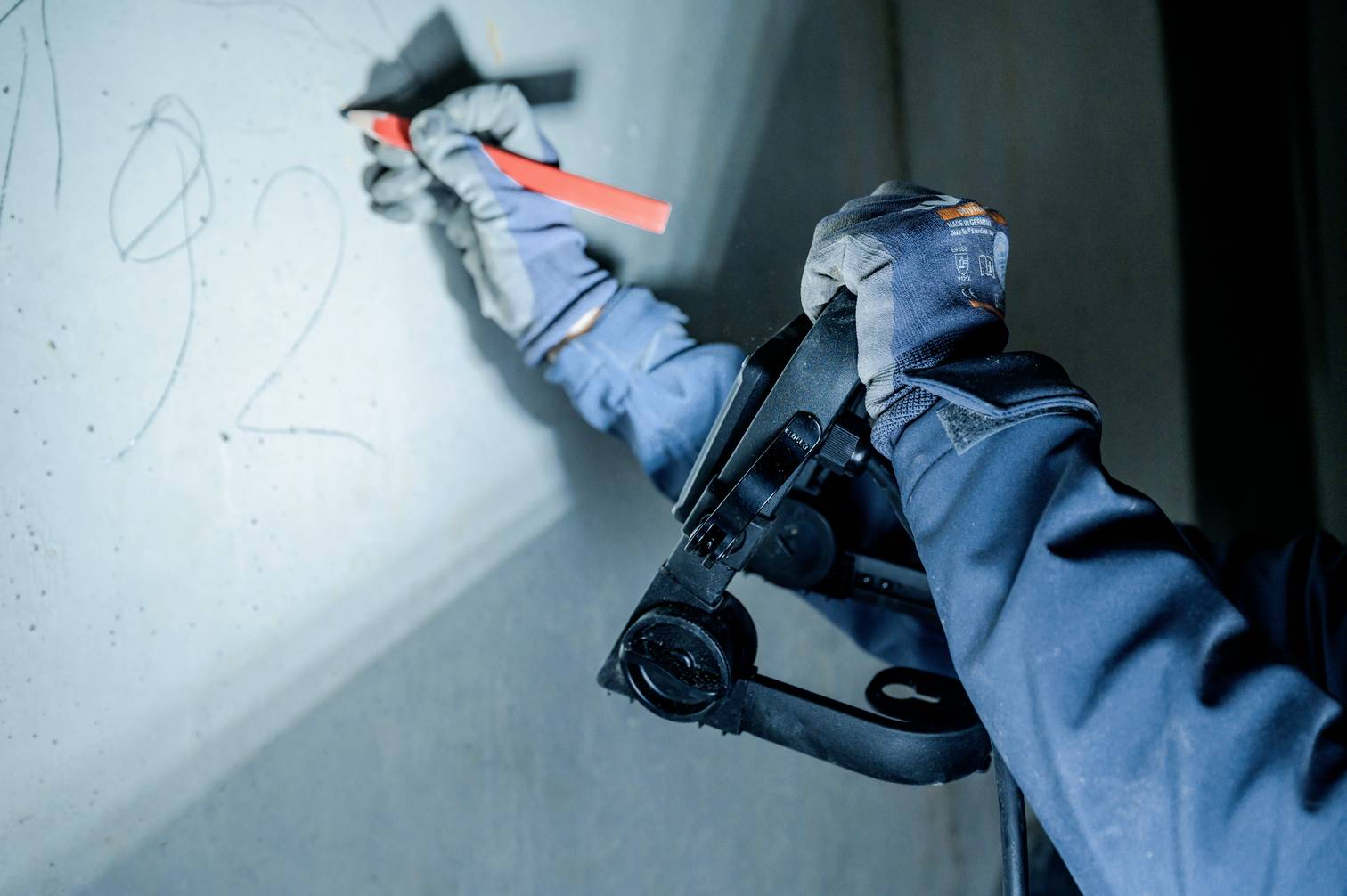 Eine Person mit Handschuhen hält ein Messgerät und zeichnet Linien an eine Wand. Das Licht betont die Handschuhe und das Werkzeug.