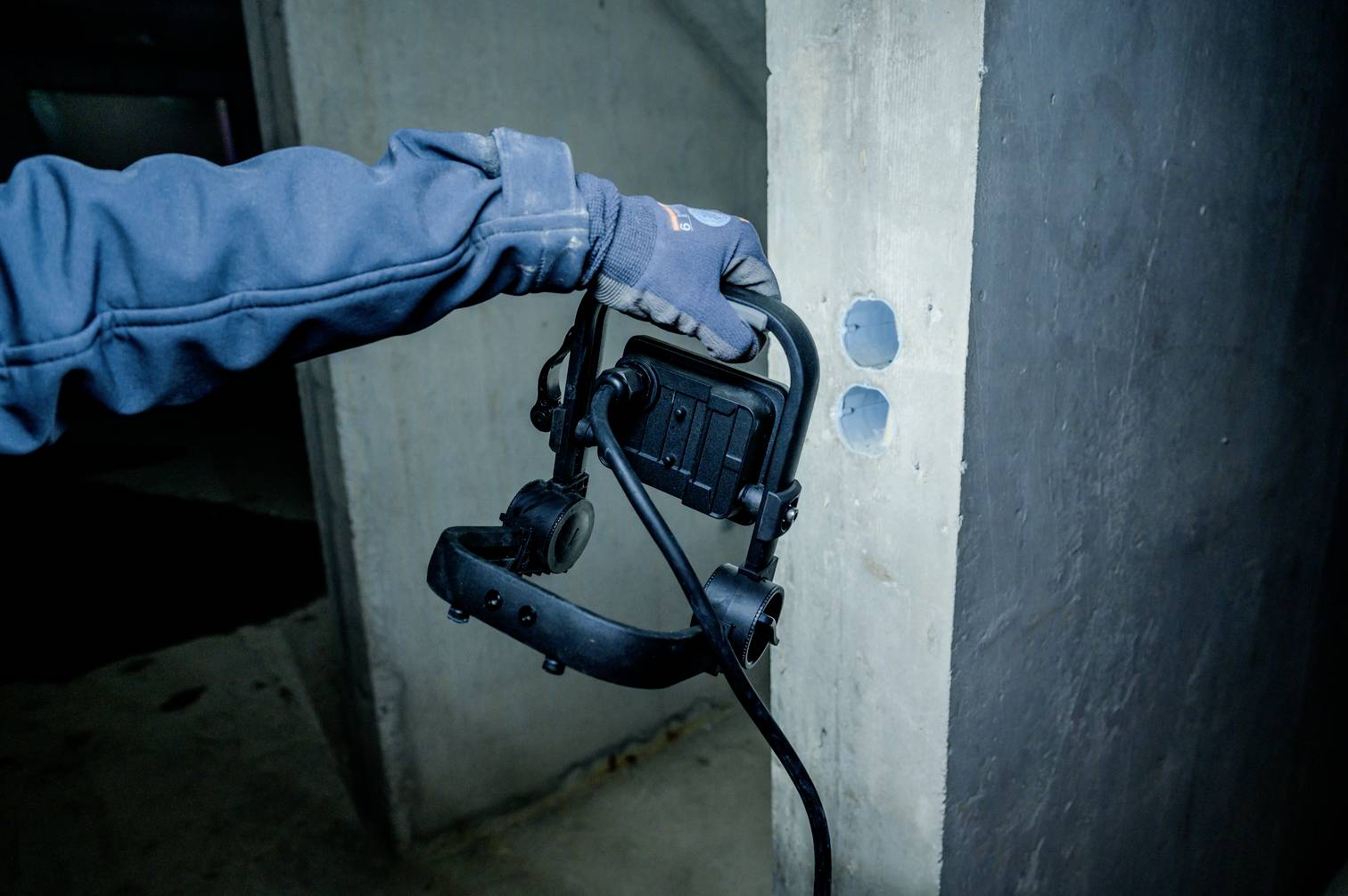Eine Person in blauer Jacke hält ein schwarzmetallisches Gerät vor eine Betonwand in einem dunklen Raum.