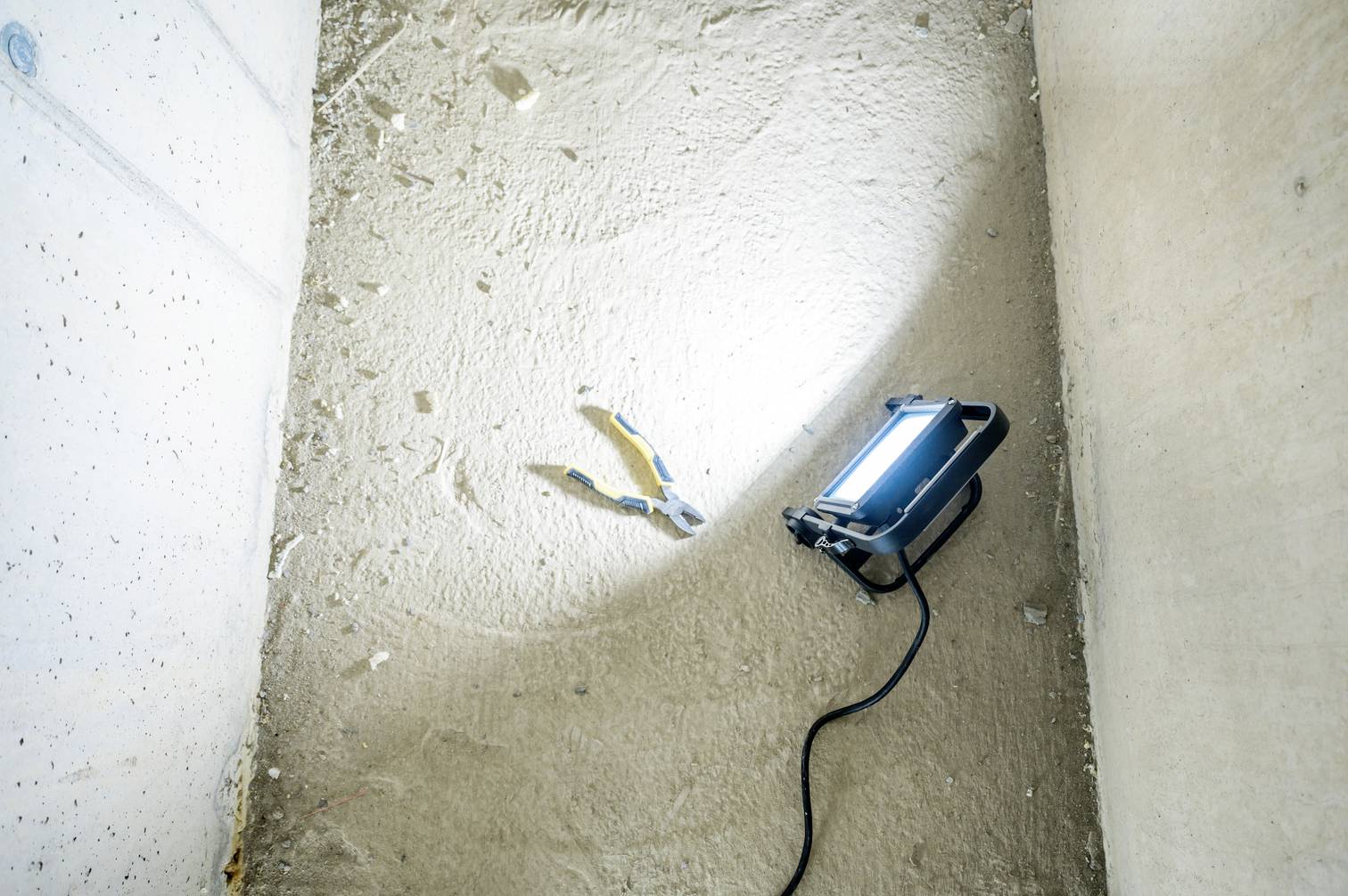 Schmutziger Boden in einem Keller mit einer Arbeitsleuchte und einer Zange. Die Leuchte wirft einen Lichtkegel auf die graue Wand.