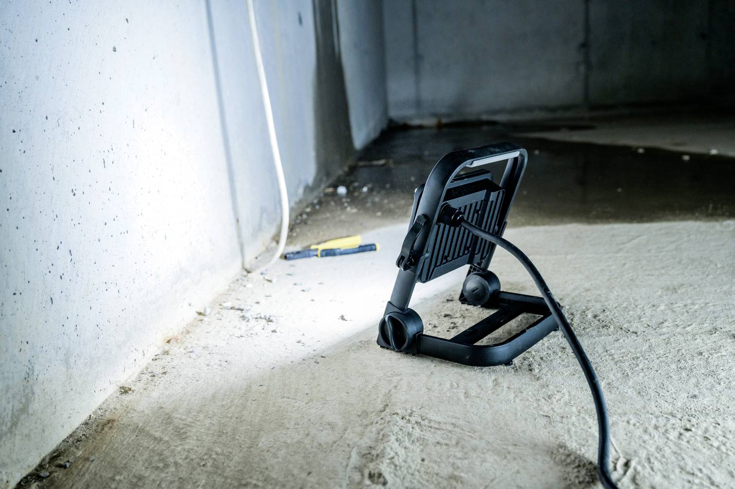 Ein Arbeitslicht liegt auf einem Betonboden in einem dunklen Raum, beleuchtet eine Ecke. Ein blau-gelber Kugelschreiber liegt daneben.