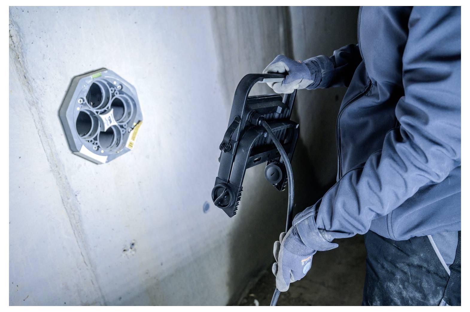 Arbeiter in Handschuhen und blauem Overall hält ein elektrisches Kabel und ein schwarzes Rohrzug-Werkzeug neben einer wandmontier Ausschalterdose während der Installation oder Reparatur.