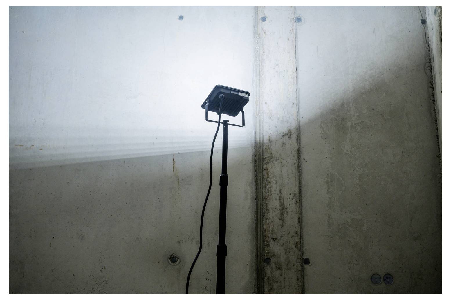 Ein tragbares Arbeitslicht auf einem Ständer beleuchtet eine kahle Betonwand und eine Ecke in einem dunklen Raum, wobei ein Kabel nach unten hängt.