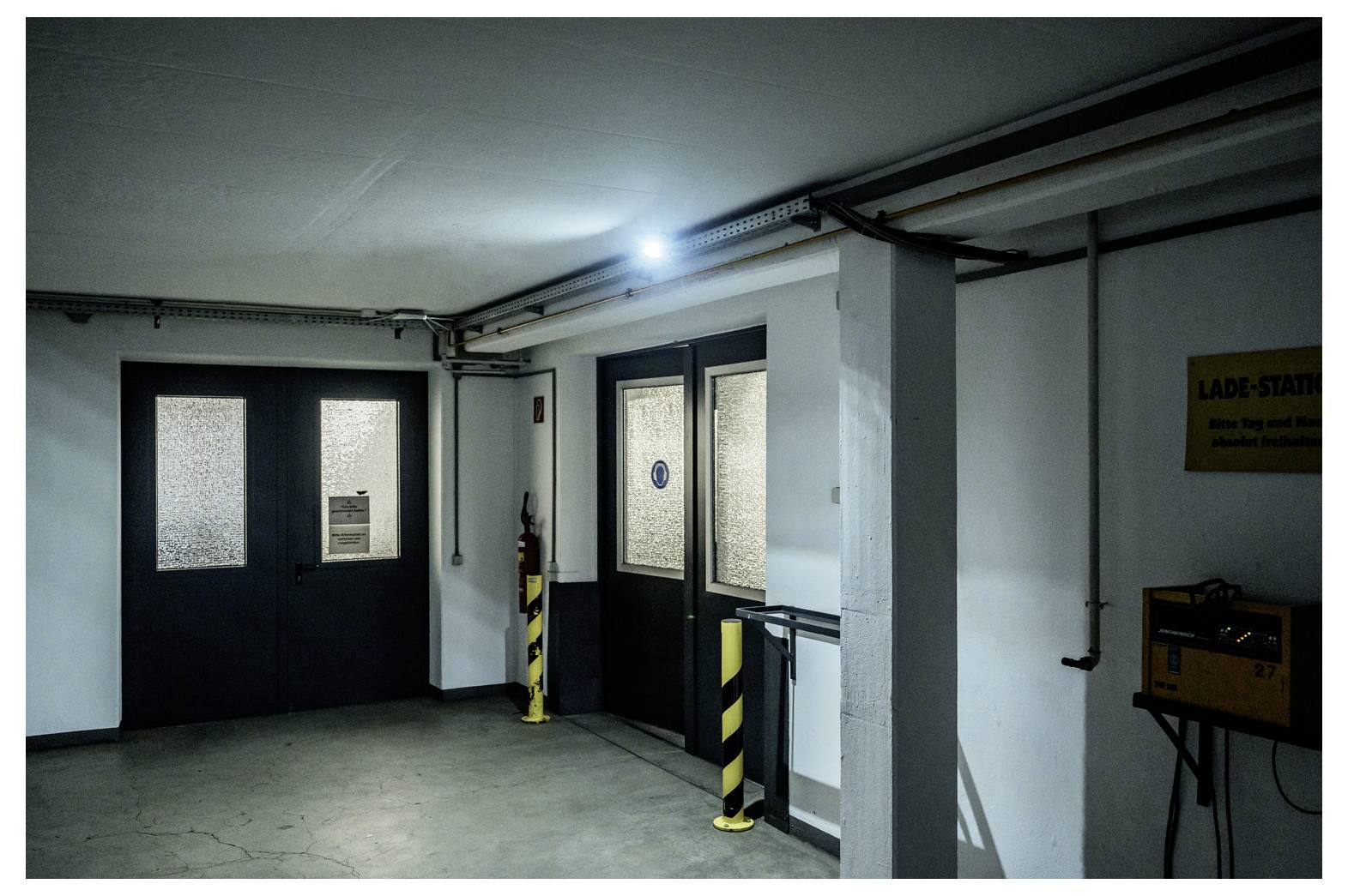 Düsterer Eingang zu einem unterirdischen Parkhaus mit schwarzen Doppeltüren, einer Seitenpedestrantür und gestreiften Sicherheitspollern