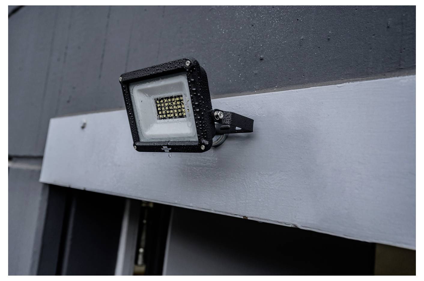 Schwarzer LED-Flutlichtstrahler auf einer dunklen Außenwand über einem silbernen Metallpaneel montiert.