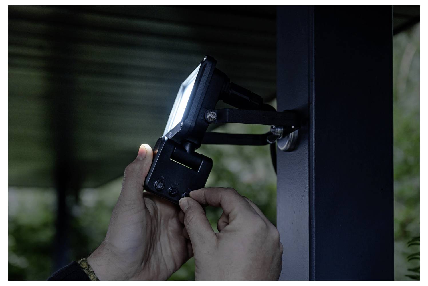 Hände stellen ein montiertes Außensicherheitslicht auf einem Metallpfosten ein, wobei die Lampe im Tageslicht nach links ausgerichtet ist.