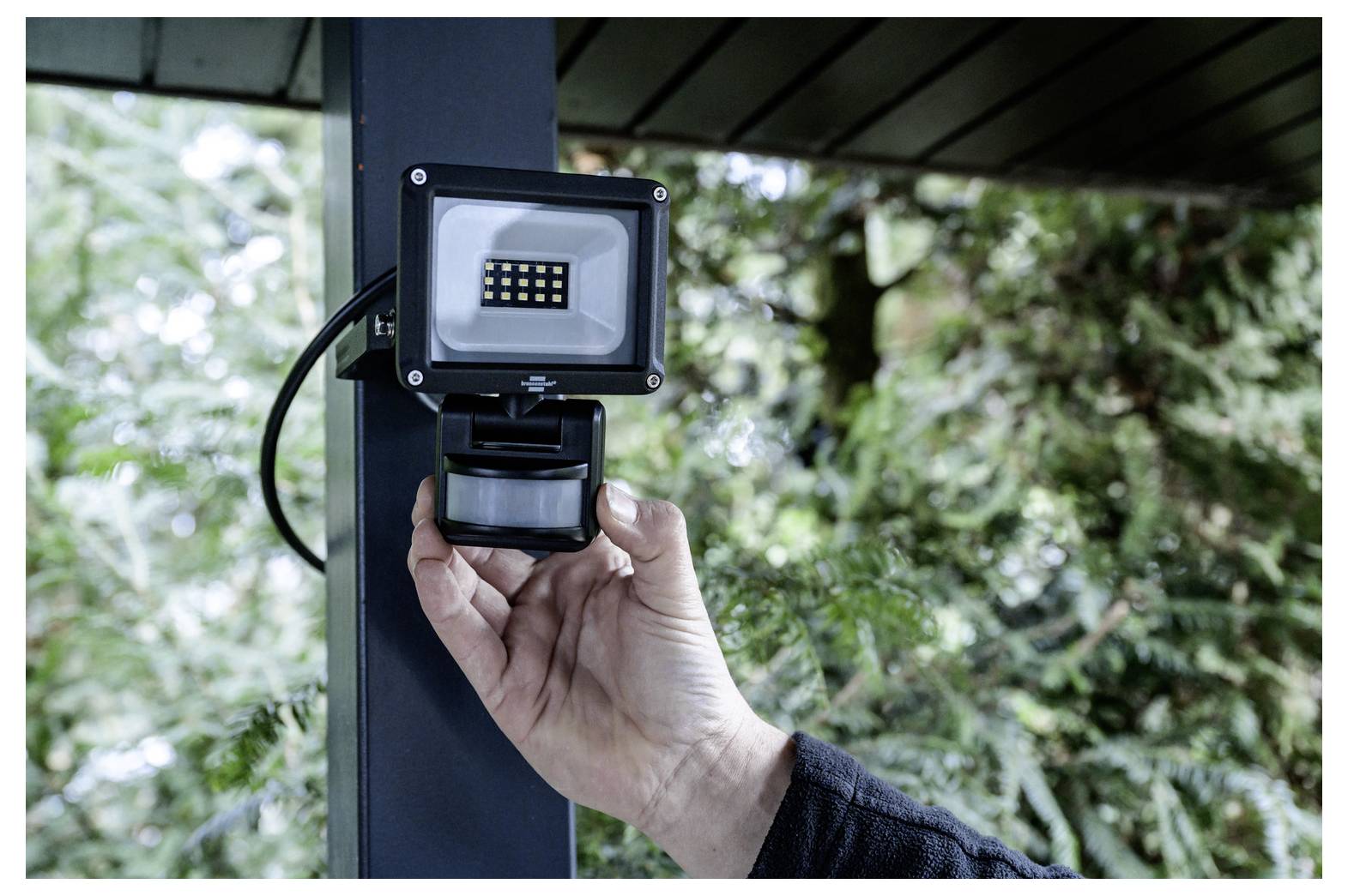 Hand adjusting a motion sensor floodlight mounted on a porch post outdoors.<br><br>Eine Hand stellt einen Bewegungsmelder-Strahler an, der auf einem Veranda-Pfosten im Freien montiert ist.