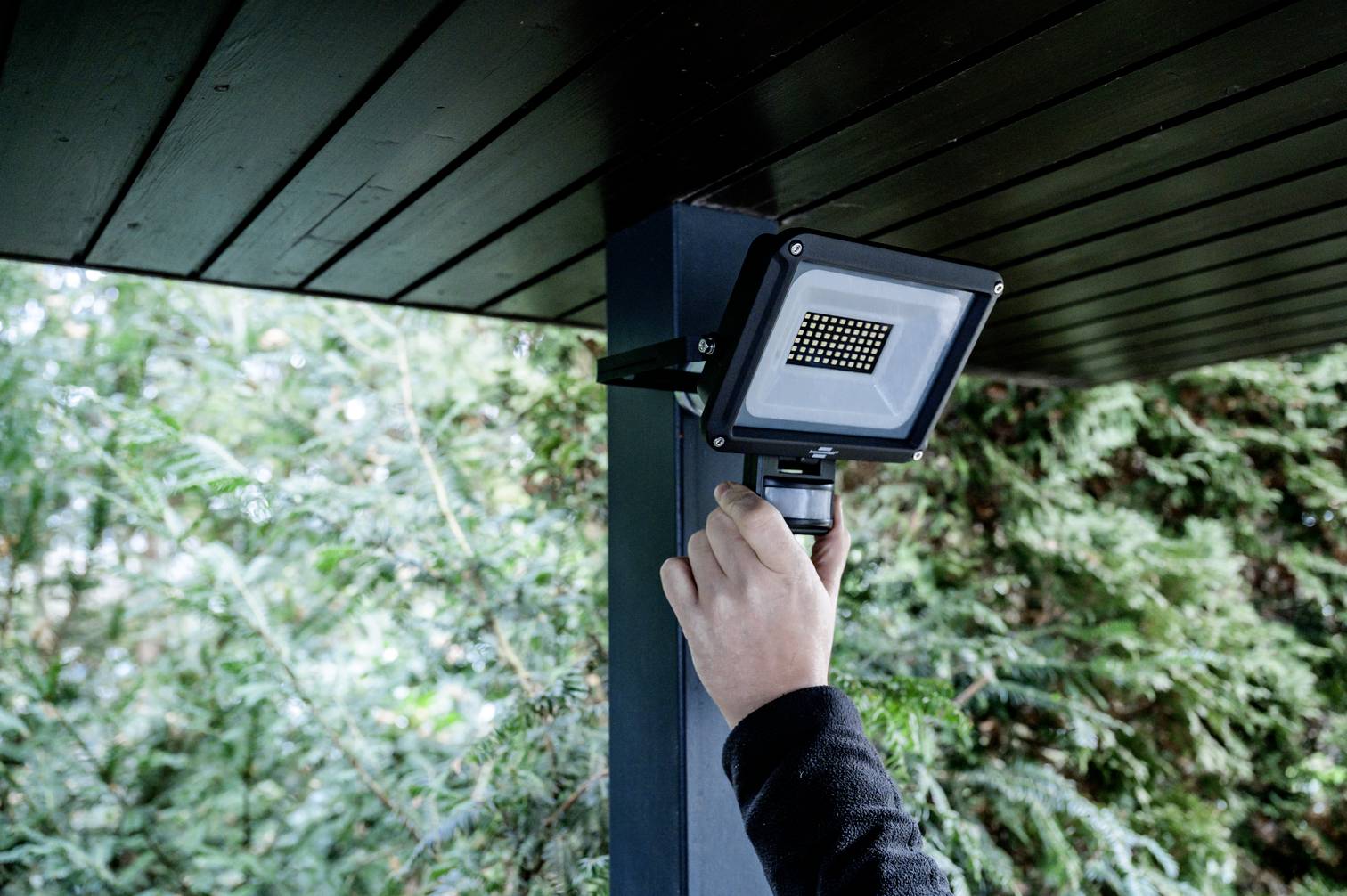 Eine Person montiert einen LED-Fluter an einem Pfosten unter einem Überhang. Im Hintergrund sind grüne Bäume sichtbar.