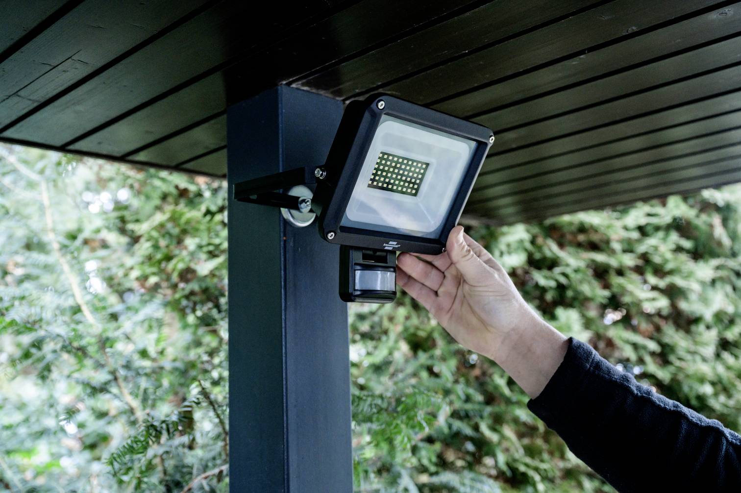 Eine Person installiert einen LED-Strahler mit Bewegungssensor an einem Holzpfosten unter einem Vordach im Freien.