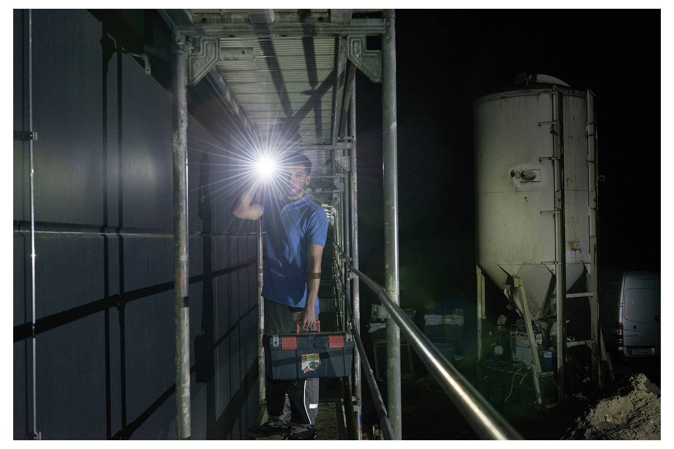 Arbeiter, der nachts auf einem Gerüst steht, eine helle Taschenlampe in der Hand hält, in einem Industriegebiet neben einem großen Tank und geparkten Fahrzeugen.