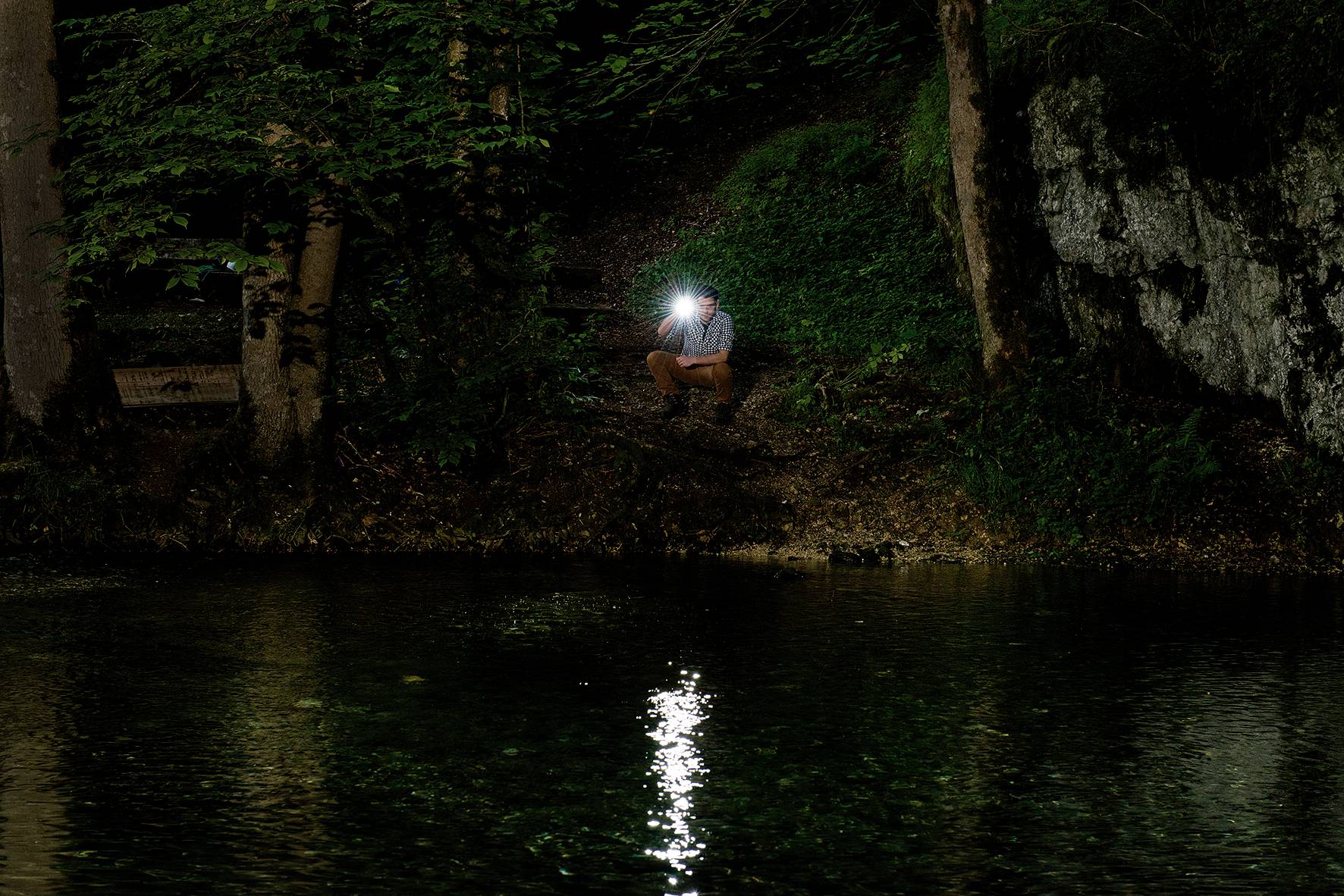 Eine Person sitzt nachts am Ufer eines Sees, hält eine Taschenlampe, die ins Wasser leuchtet. Umgeben von Bäumen und Felsen.