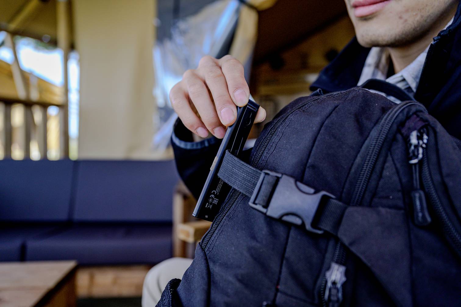 Eine Person verstaut ein schwarzes Gerät in einem schwarzen Rucksack. Im Hintergrund sind verschwommene Innenraummöbel zu sehen.