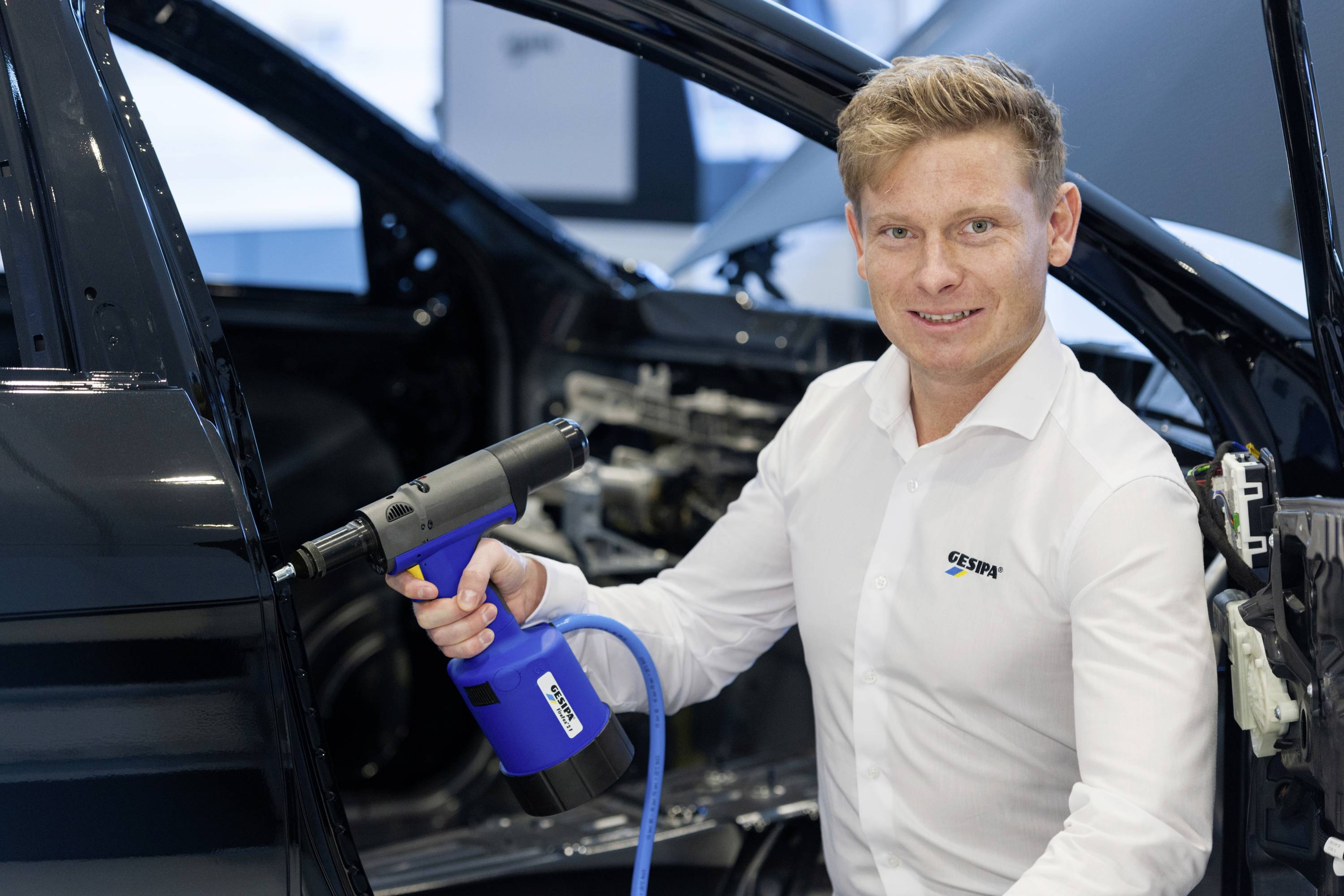 Eine Person in Arbeitskleidung hält ein Werkzeug in der Hand und steht neben einem teilweise montierten schwarzen Auto in einer Werkstattumgebung.