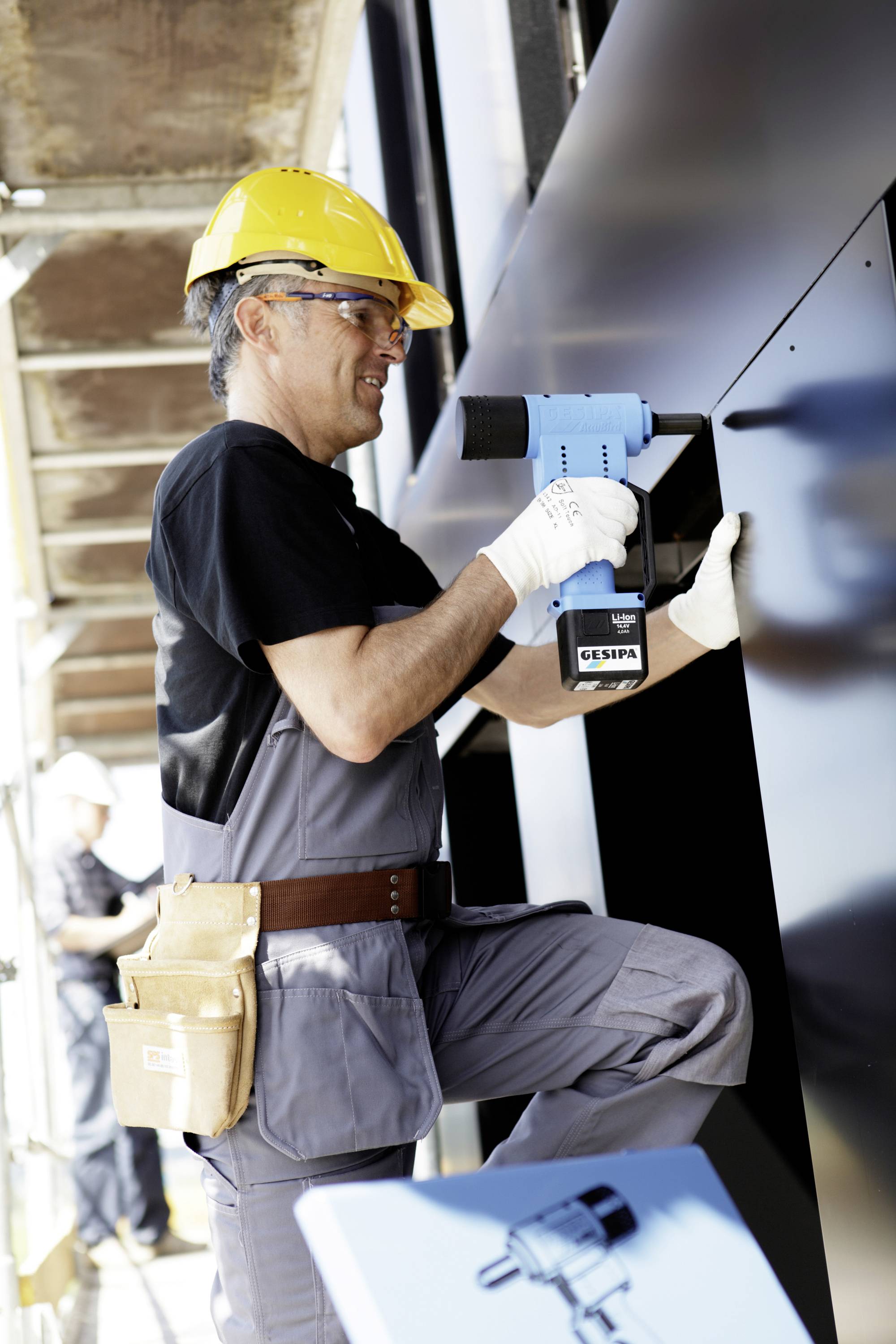 Ein Bauarbeiter in blauer Arbeitskleidung und gelbem Helm benutzt eine Bohrmaschine, um eine Metallplatte an eine Wand zu befestigen.
