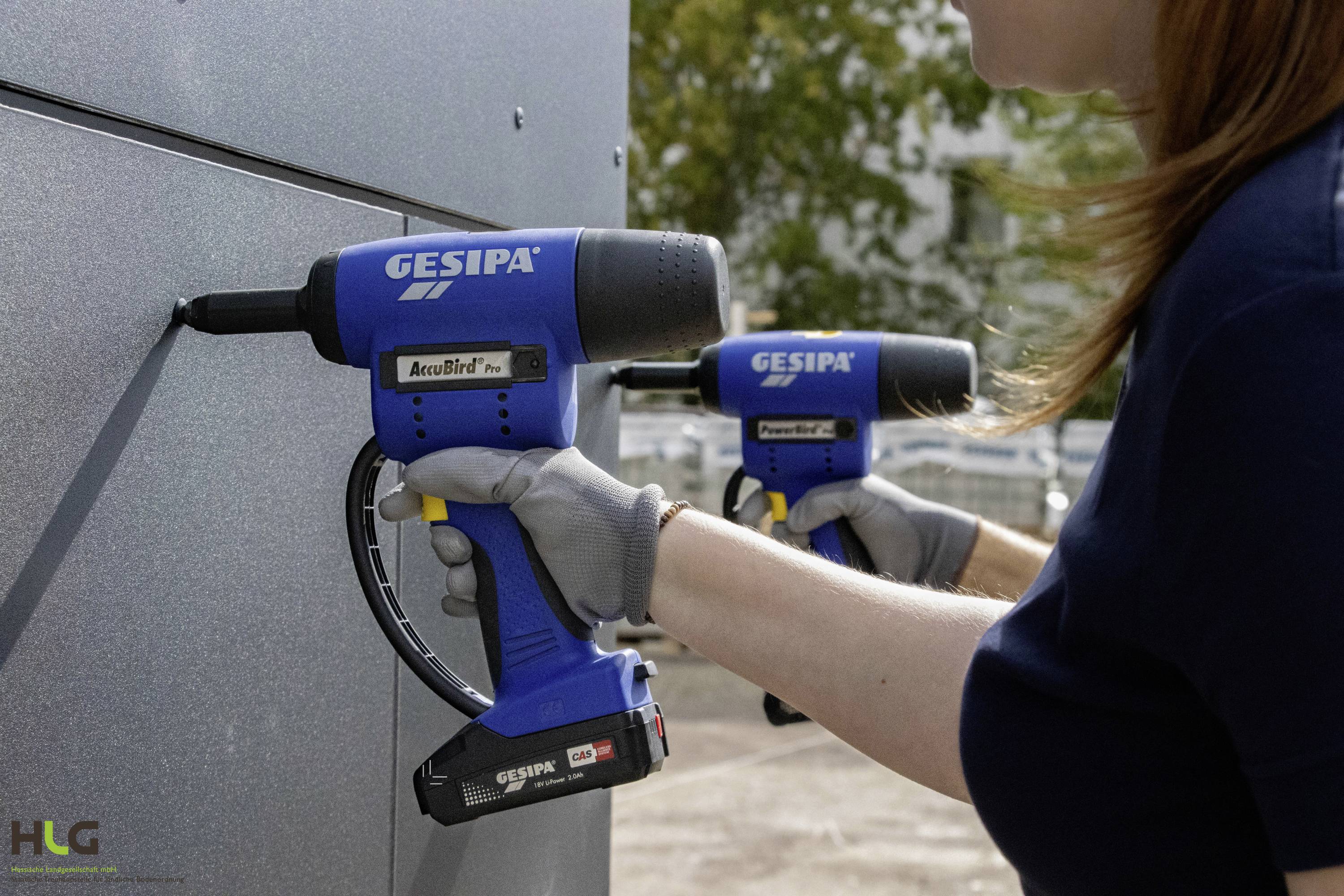 Person verwendet eine blaue Akku-Blindnietpistole an einer Metallplatte. Im Hintergrund eine zweite, unscharfe Person mit ähnlichem Werkzeug.