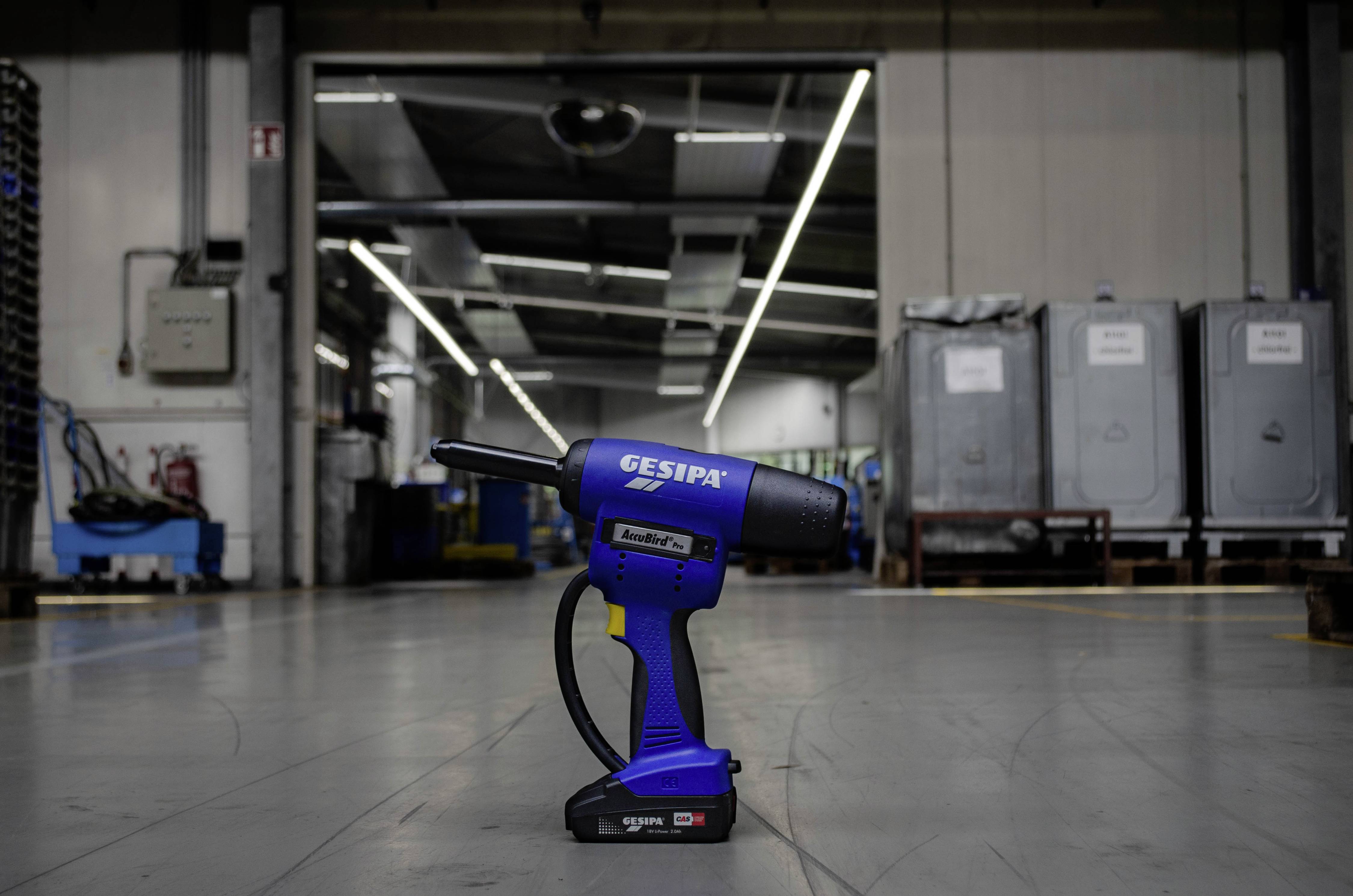 Ein blauer, akkubetriebener Blindnietwerkzeug liegt auf dem Boden einer großen, industriellen Werkhalle mit Maschinen im Hintergrund.