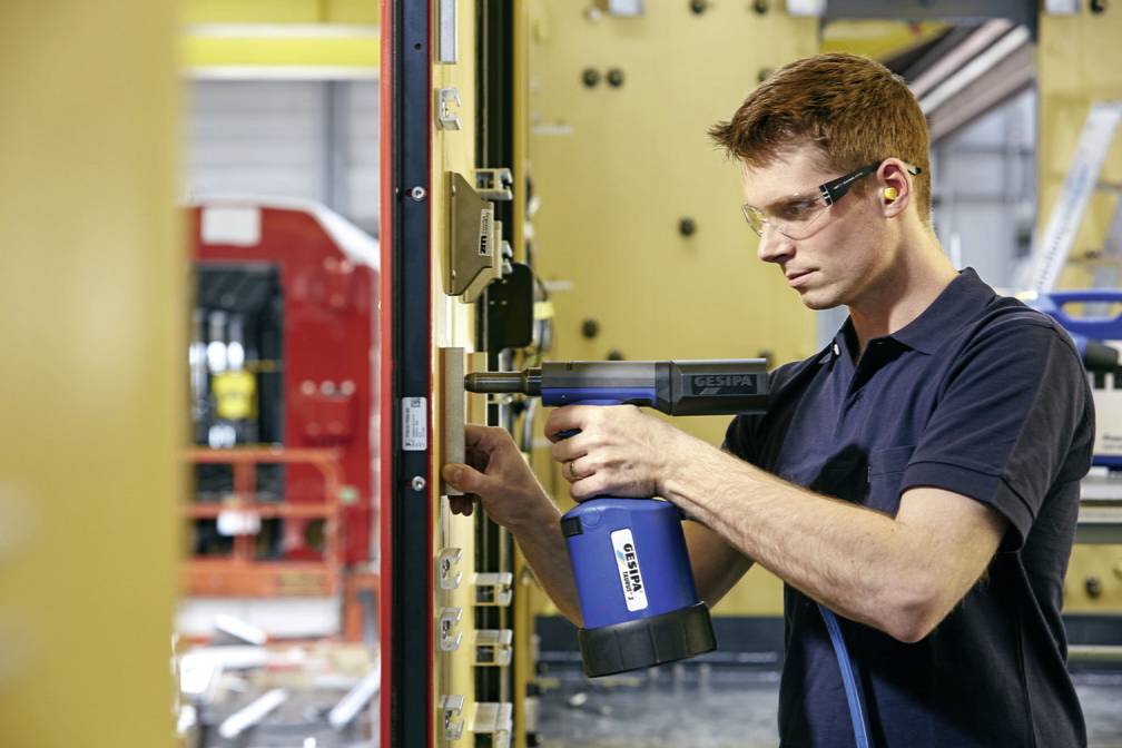 Ein junger Mann in Schutzbrille und Gehörschutz verwendet ein Elektrowerkzeug, um an einer Industriemaschine in einer Fabrikumgebung zu arbeiten.