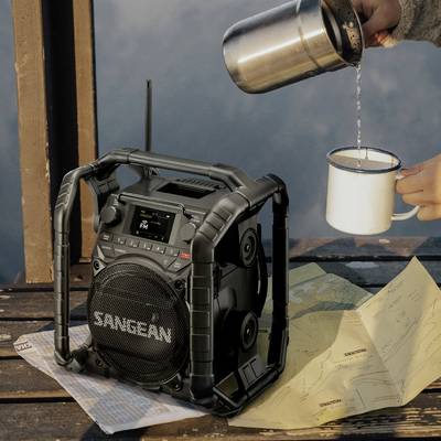 Eine Hand gießt Kaffee in eine Emaille-Tasse neben einem Radio. Karten liegen auf dem Holztisch, was auf ein Outdoor-Szenario hindeutet.