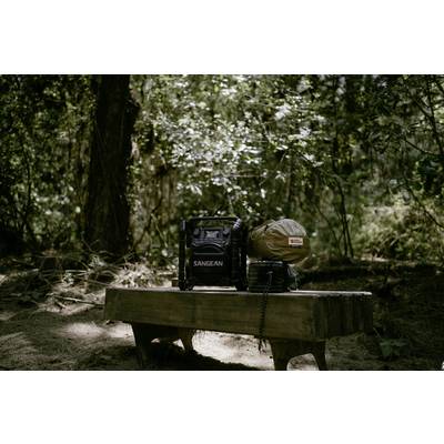 Ein Radio und eine Tasche stehen auf einer Holzbank im Wald. Sonnenlicht scheint durch die Bäume.