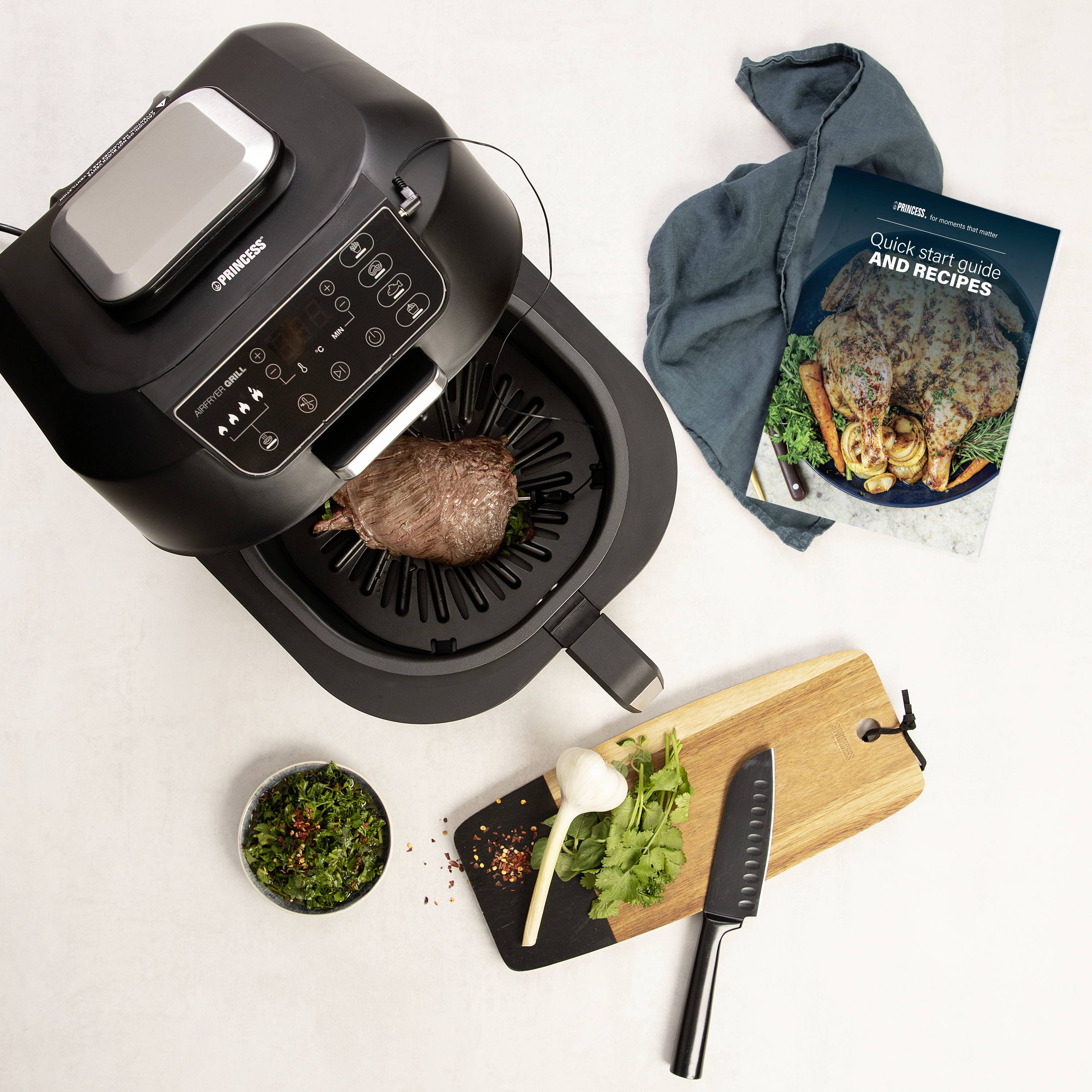 Ein Luftfritteuse mit einem Braten auf dem Grillrost. Daneben liegen ein Schneidebrett mit Messer, Knoblauch, Kräutern und ein Rezeptheft.
