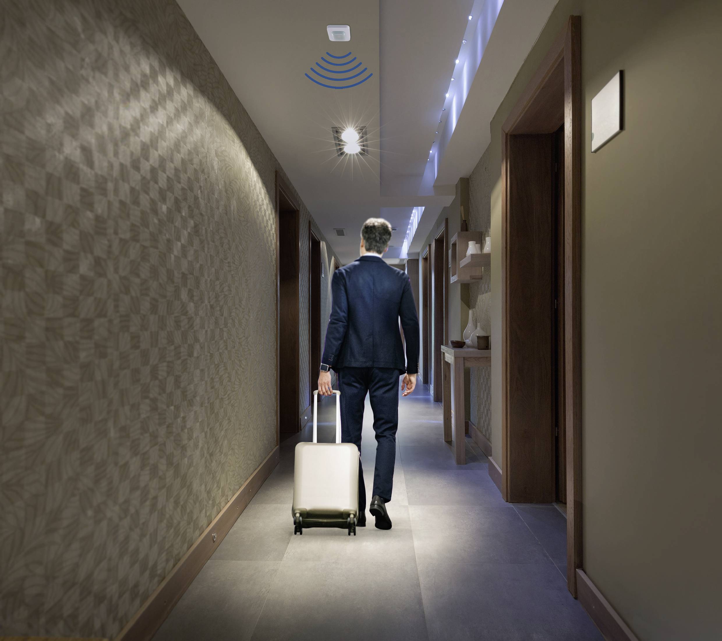 Eine Person in einem Anzug zieht einen Rollkoffer durch einen beleuchteten Hotelflur.