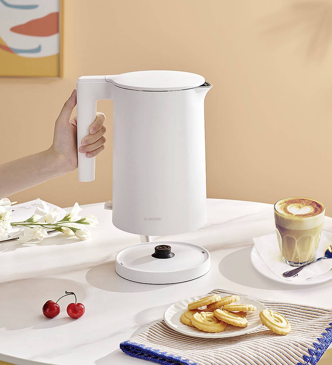 Eine Person hält einen weißen Wasserkocher über einem Tisch mit Keksen, einer Kaffeetasse und Kirschen. Im Hintergrund ist ein buntes Bild.