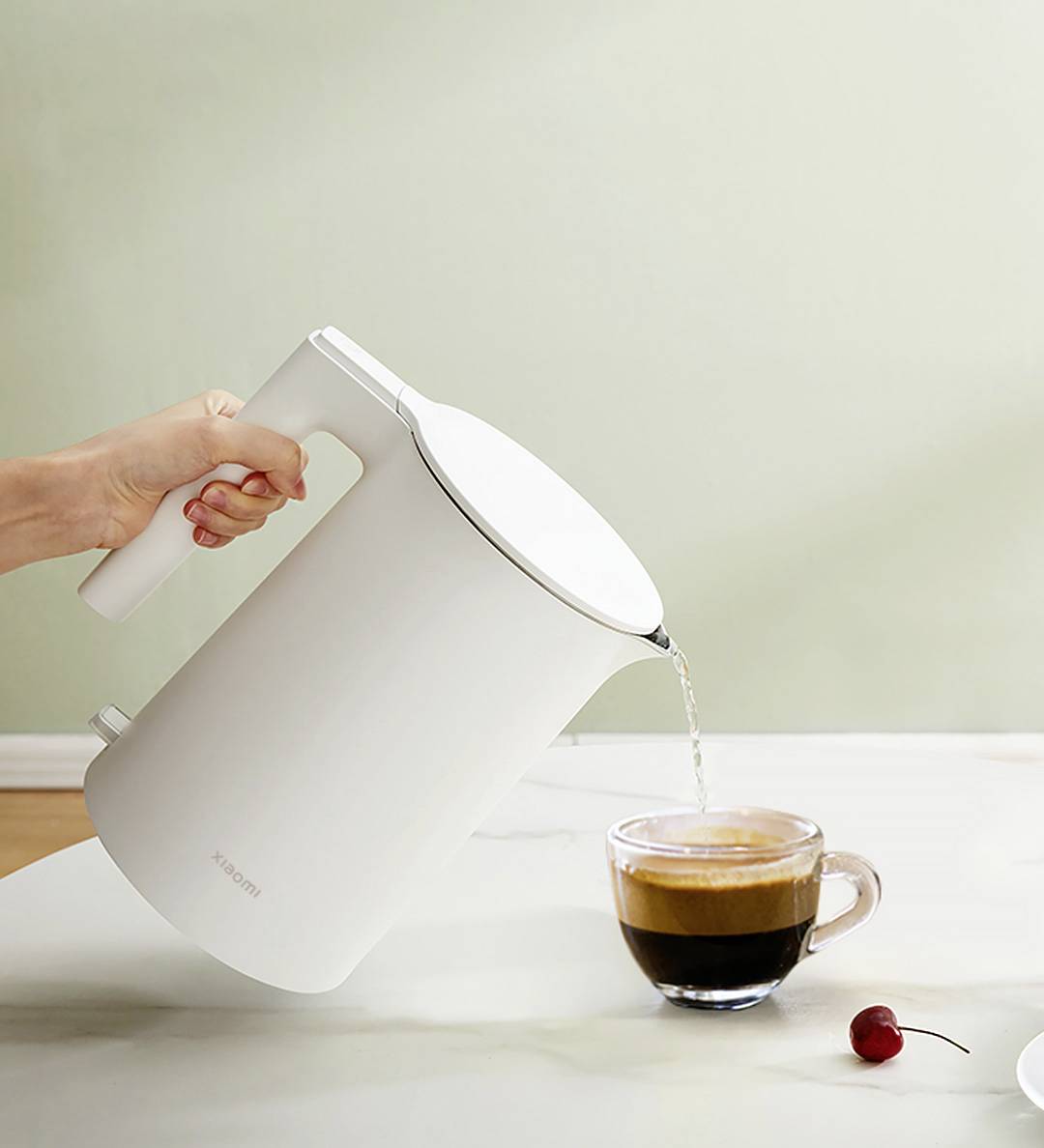 Eine helle Kanne gießt heißes Wasser in eine Tasse Kaffee auf einem Tisch. Daneben liegt eine Kirsche.