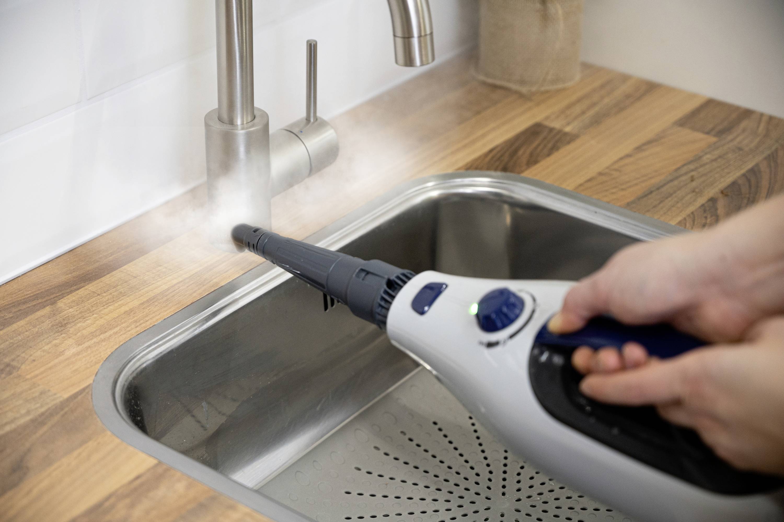 Eine Person reinigt mit einem Dampfreiniger den Wasserhahn eines Spülbeckens, dabei entsteht sichtbarer Dampf um den Auslauf.
