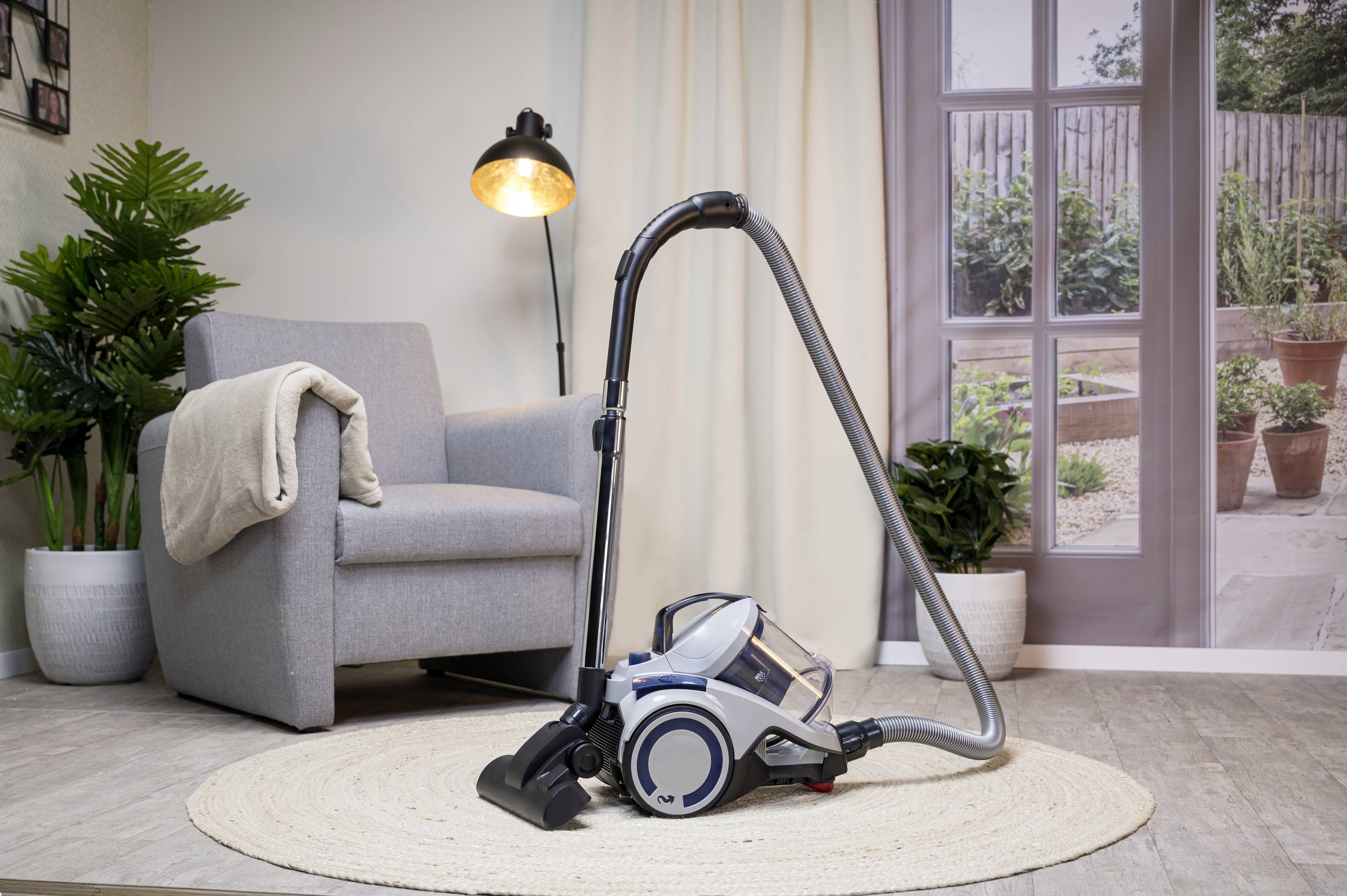 Staubsauger im Wohnzimmer auf Teppich neben grauem Sessel, Stehlampe und Fenster mit Blick auf Gartenterrasse.