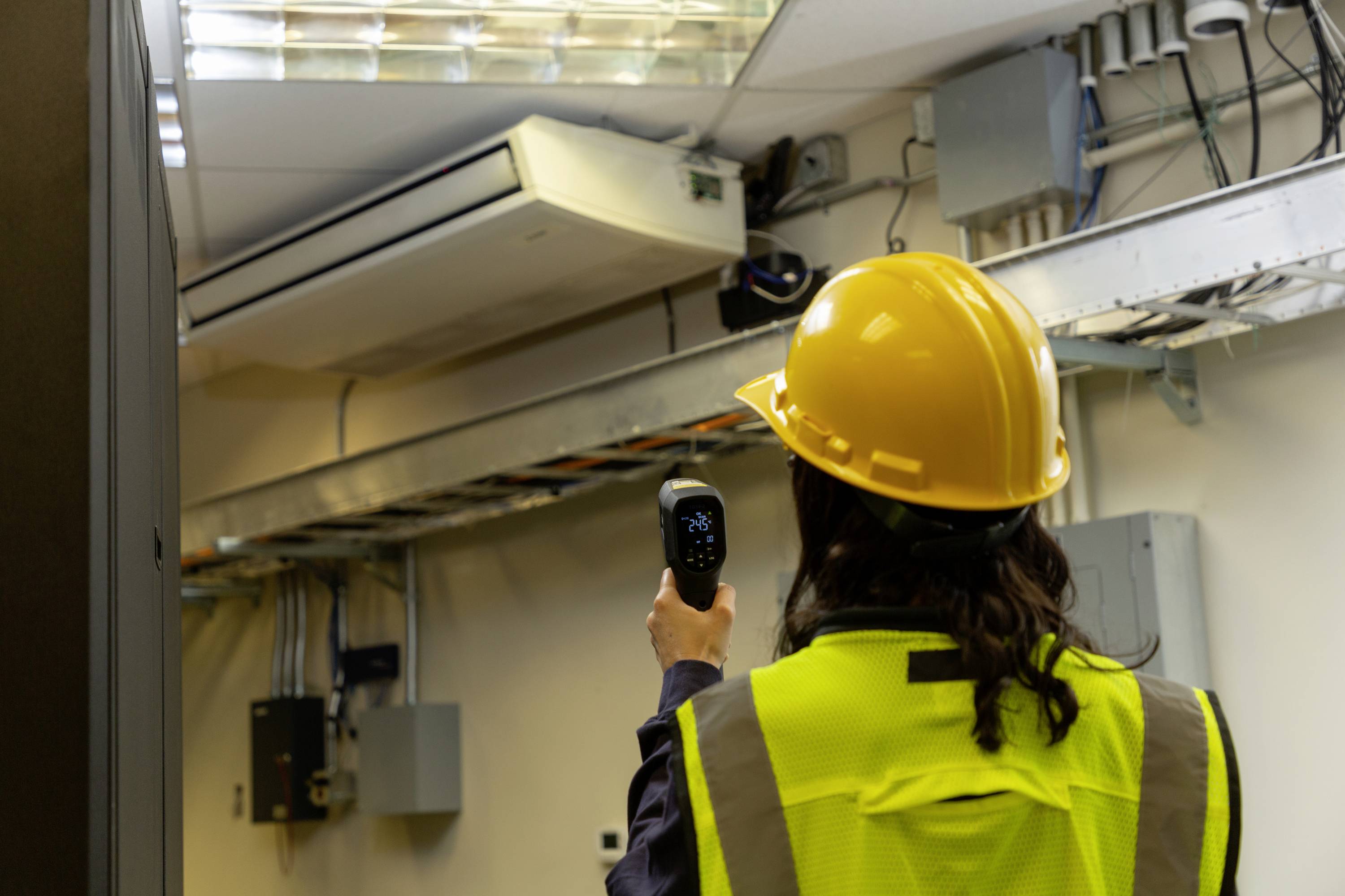 Eine Person mit Helm und Warnweste prüft mit einem Handgerät die Temperatur in einem Serverraum. Klimaanlage an der Wand.