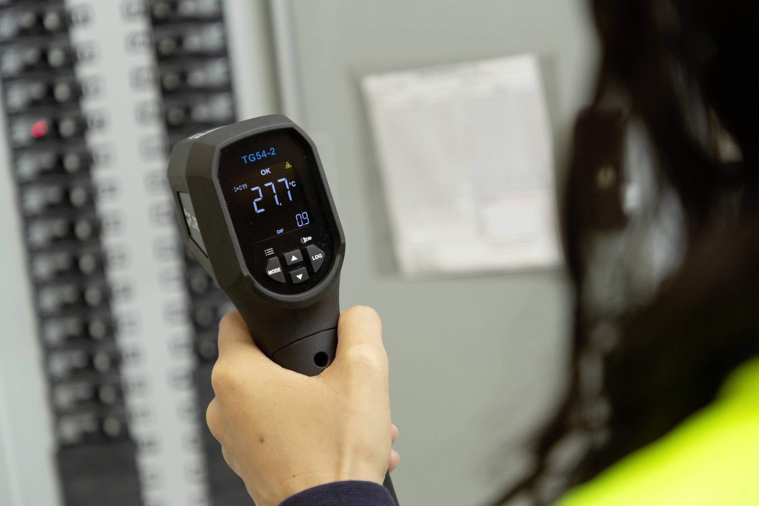 Eine Person hält einen Infrarot-Thermometer, der eine Temperatur von 27,7 Grad Celsius zeigt, vor einem Schaltschrank.