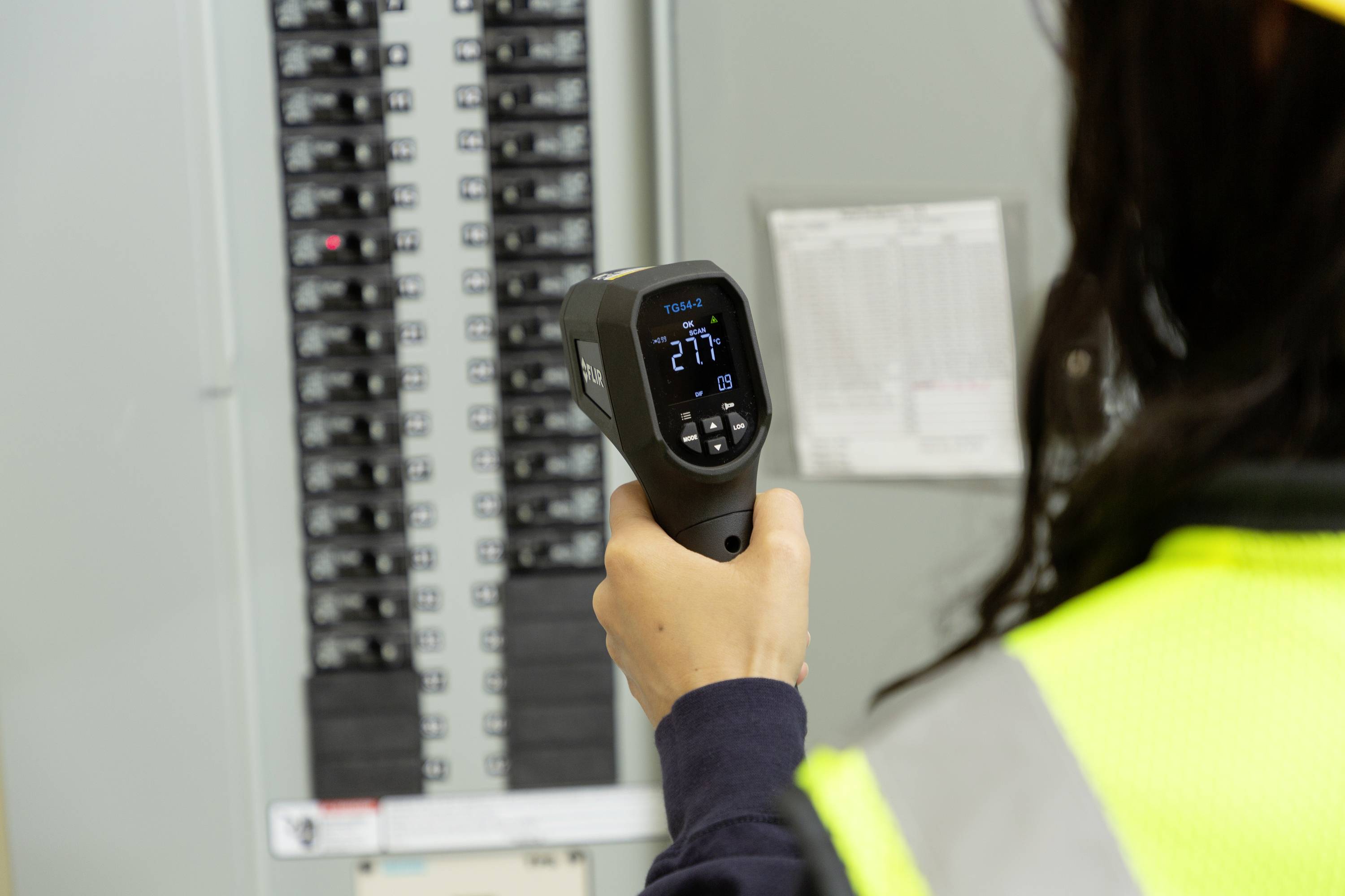 'Person misst Temperatur an einem Schaltschrank mit einem Infrarotthermometer. Anzeige zeigt 27,7 Grad Celsius.'