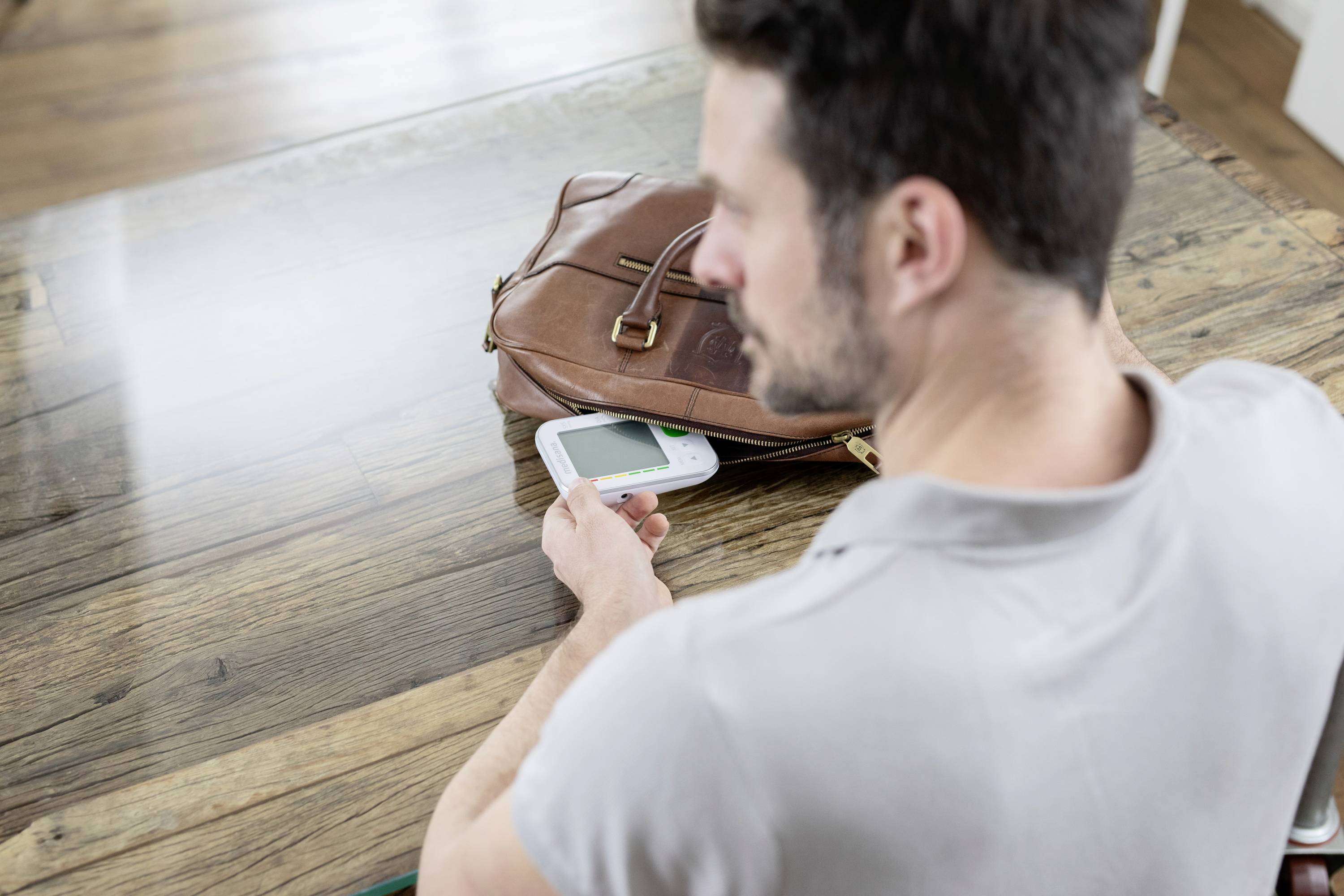 Ein Mann sitzt an einem Tisch und schaut auf ein Blutzuckermessgerät in seiner Hand. Daneben liegt eine Ledertasche.
