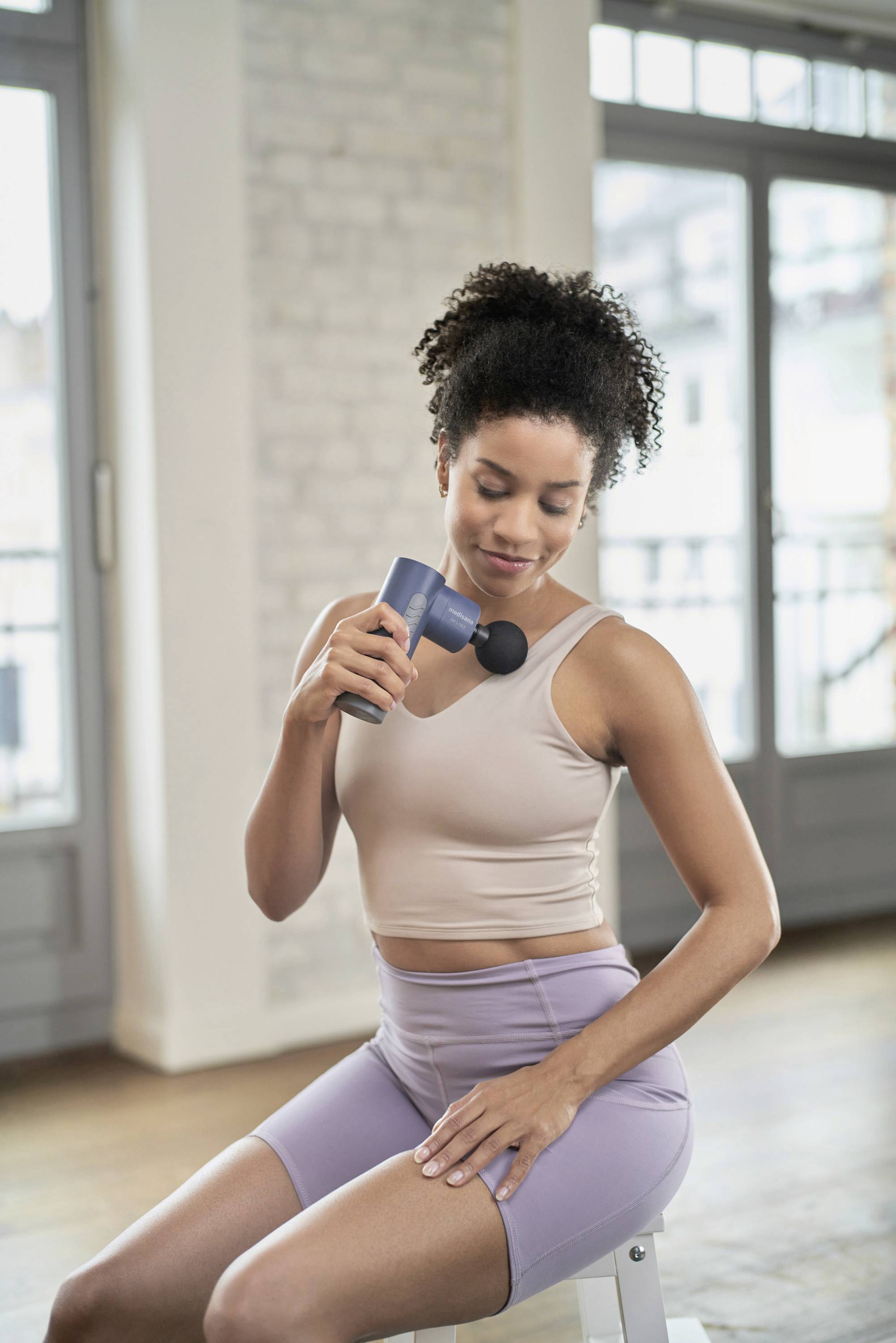 Eine Frau benutzt eine Massagepistole auf ihrer Schulter. Sie sitzt drinnen vor großen Fenstern und trägt Sportkleidung.
