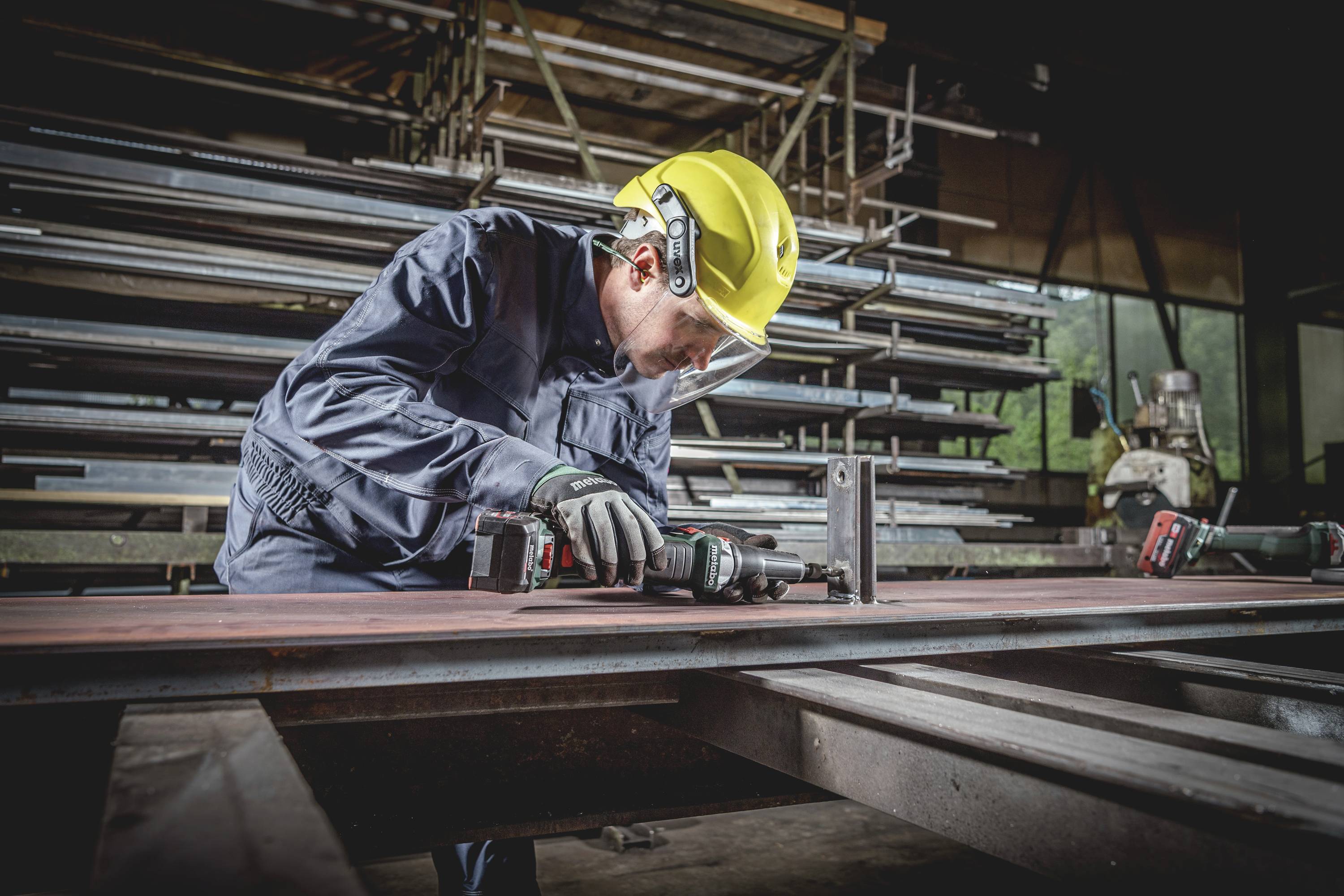 Eine Person in Arbeitskleidung und gelbem Schutzhelm bedient eine Maschine in einer Metallwerkstatt, umgeben von Stahlträgern.