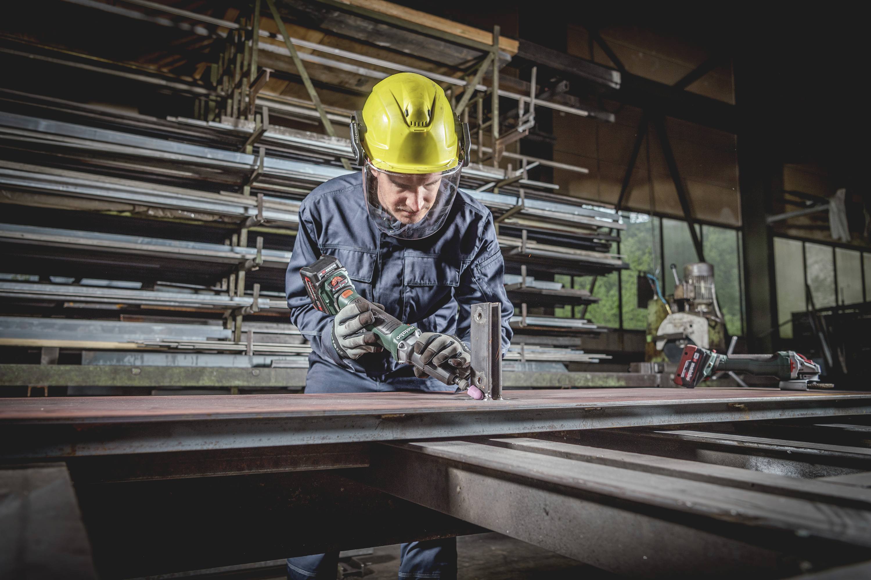 Ein Arbeiter in Schutzkleidung und gelbem Helm bedient ein Elektrowerkzeug in einer Werkstatt, um Metallteile zu bearbeiten.
