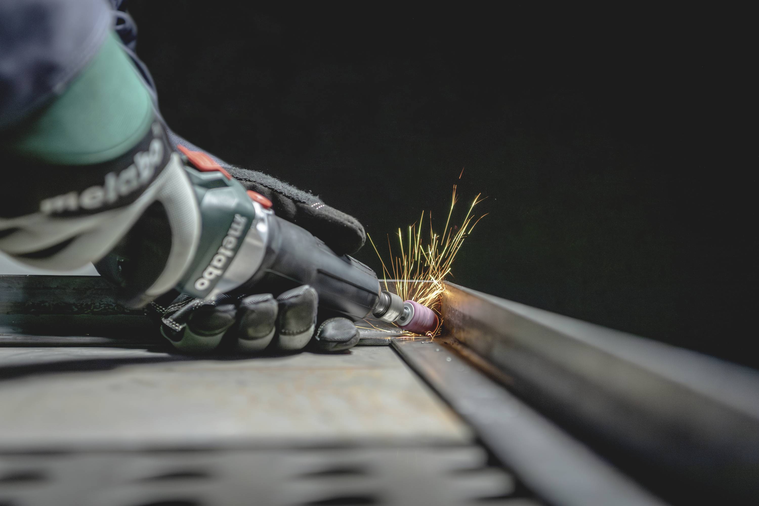 Eine Person trägt Handschuhe und schleift mit einem Winkelschleifer Metall, wobei Funken sprühen.