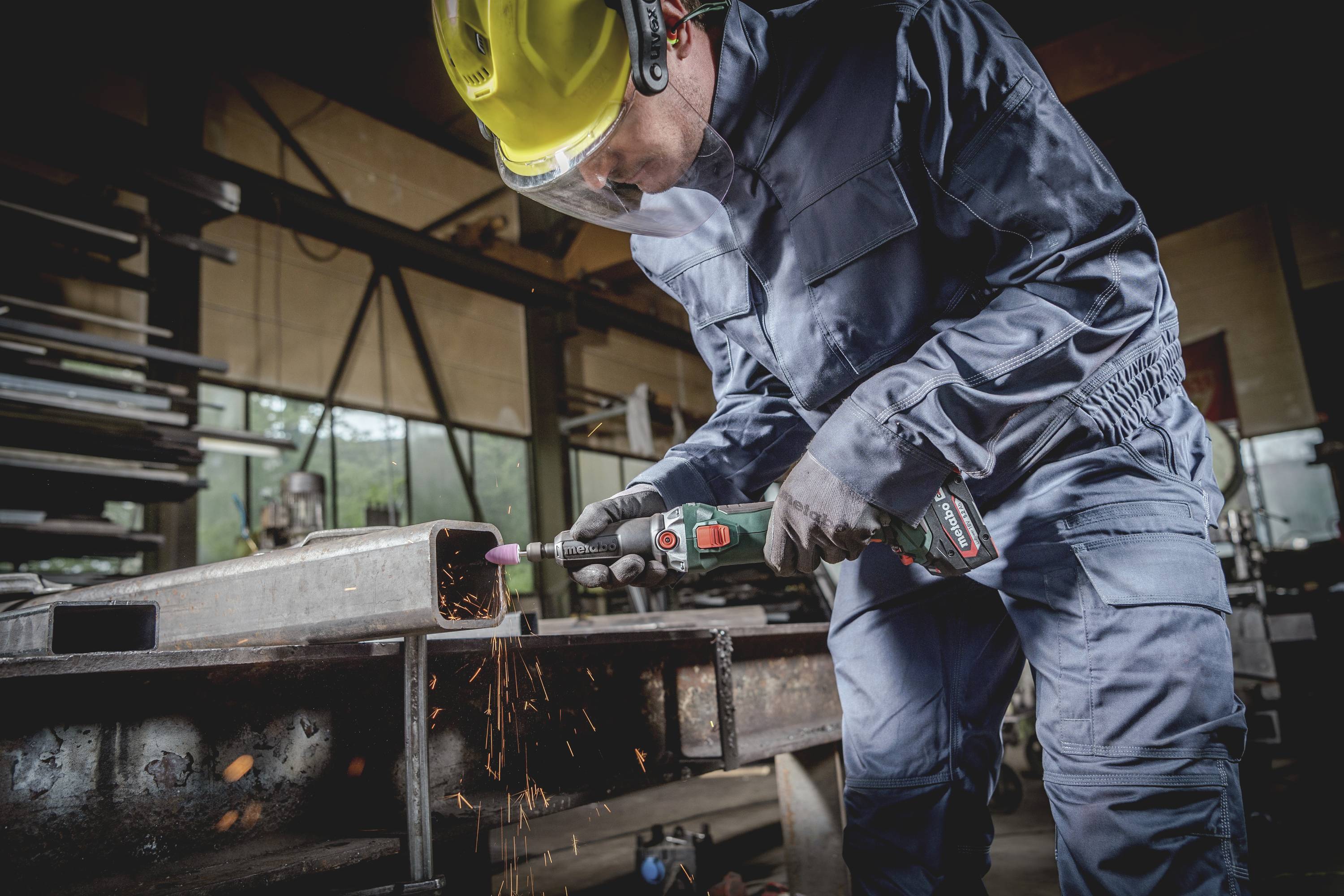 Ein Arbeiter in Schutzkleidung schleift Metall mit einem Winkelschleifer in einer Werkstatt; Funken fliegen dabei.